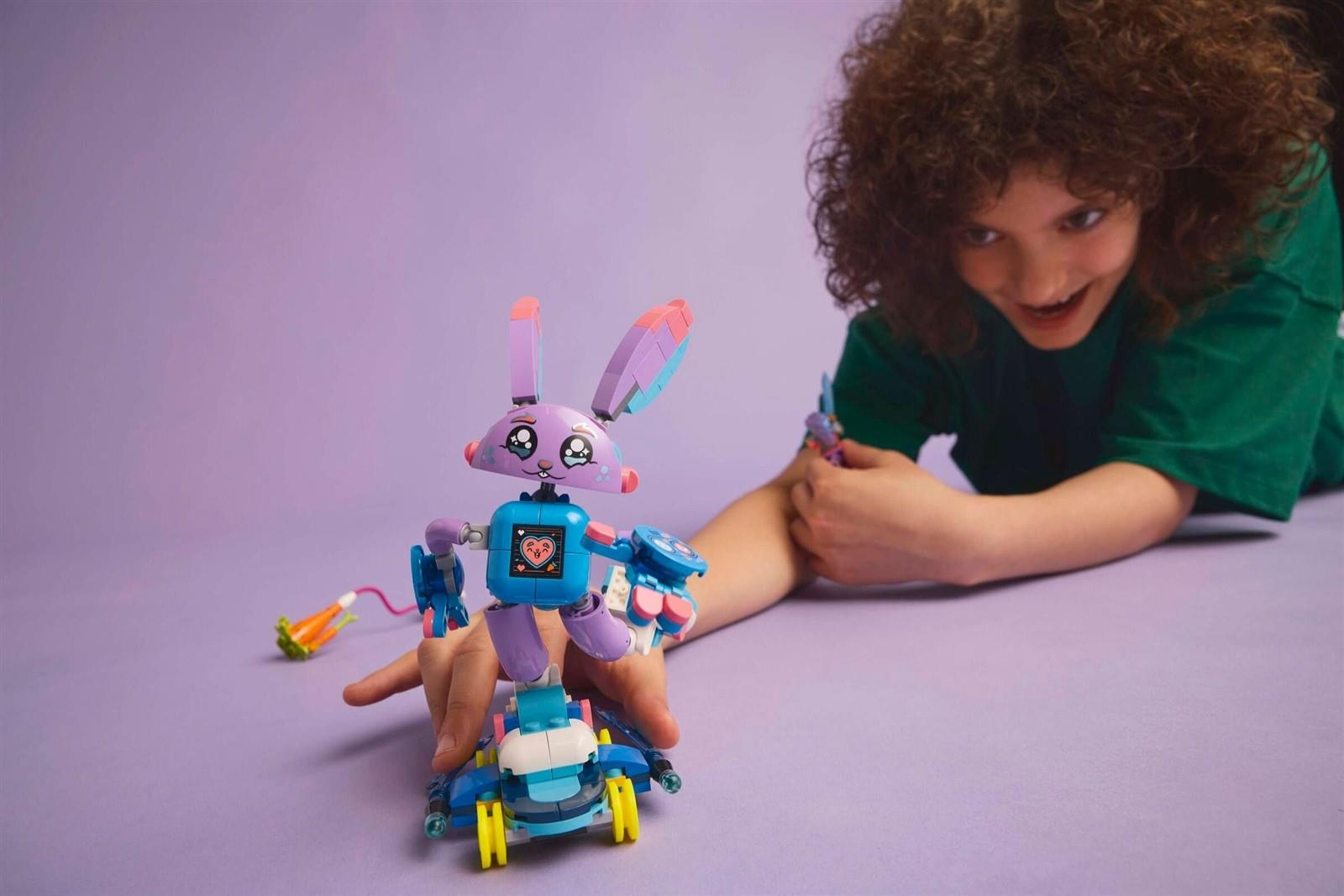 Ein Kind in grünem Shirt spielt mit einer lila Lego-Hasenfigur auf einem blauen Wagen vor lilafarbenem Hintergrund.
