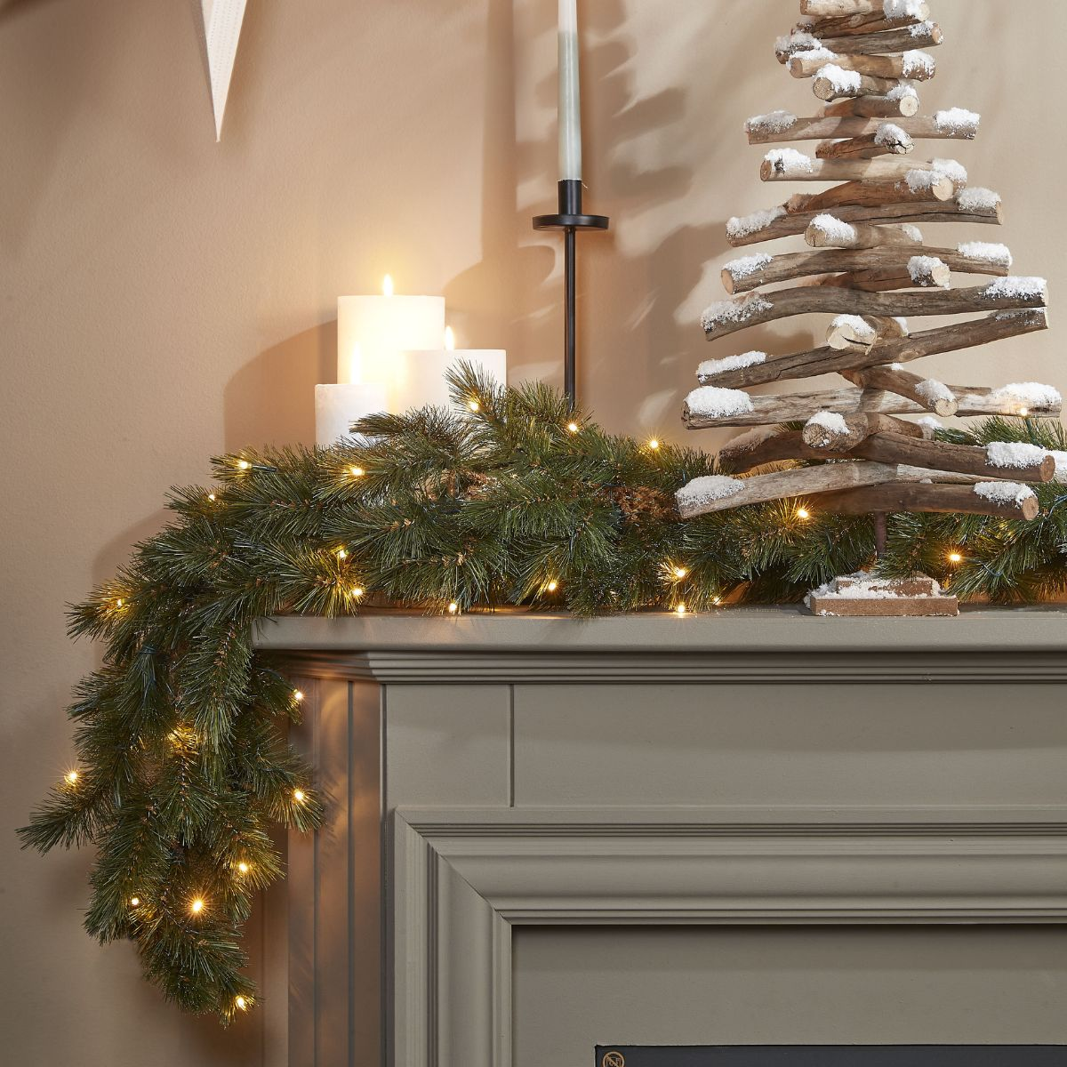 Festliche Kaminsims-Dekoration mit Tannengirlande, Lichtern, Kerzen und einem Holz-Weihnachtsbaum.