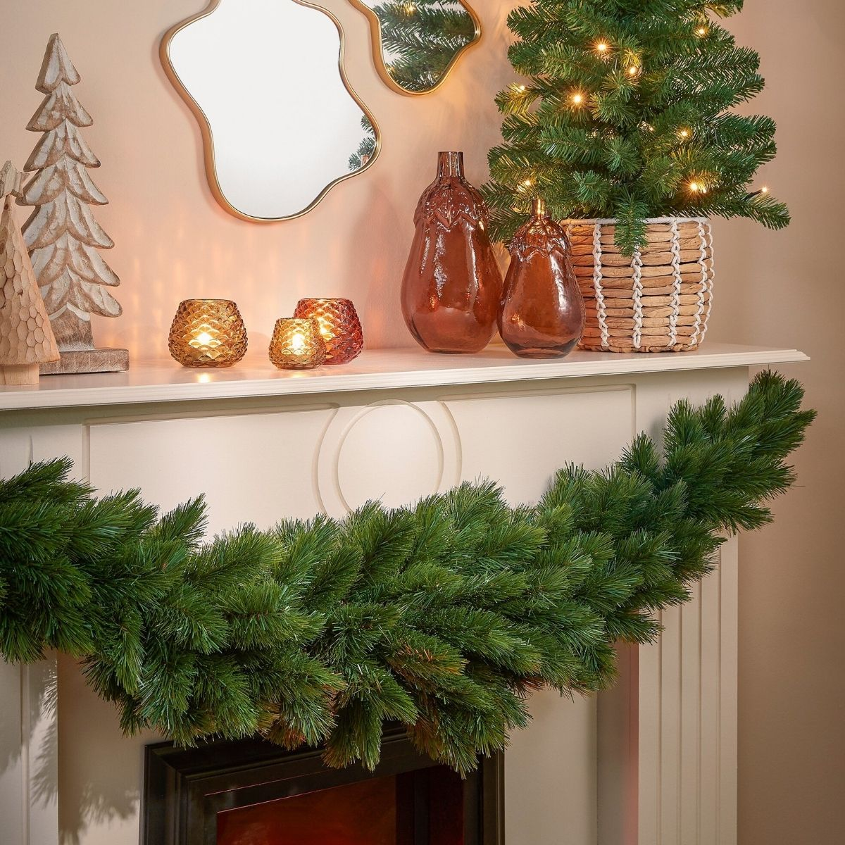 Manteau de cheminée décoré d'une guirlande de pin, de mini sapins, de vases et de bougies allumées.