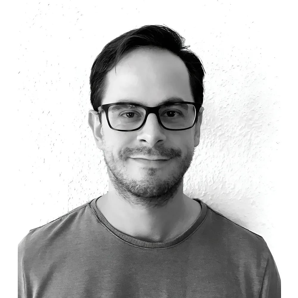 Ein Schwarz-Weiß-Porträt eines smiling Mannes mit Brille vor einer weißen Wand.