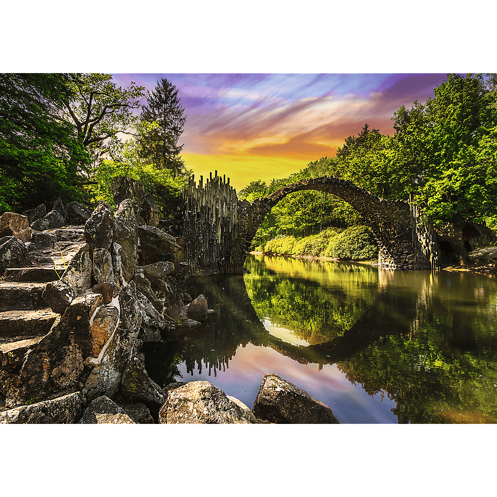 Eine steinerne Bogenbrücke spannt sich über einen Fluss, gesäumt von üppigem Grün und einem malerischen Abendhimmel.