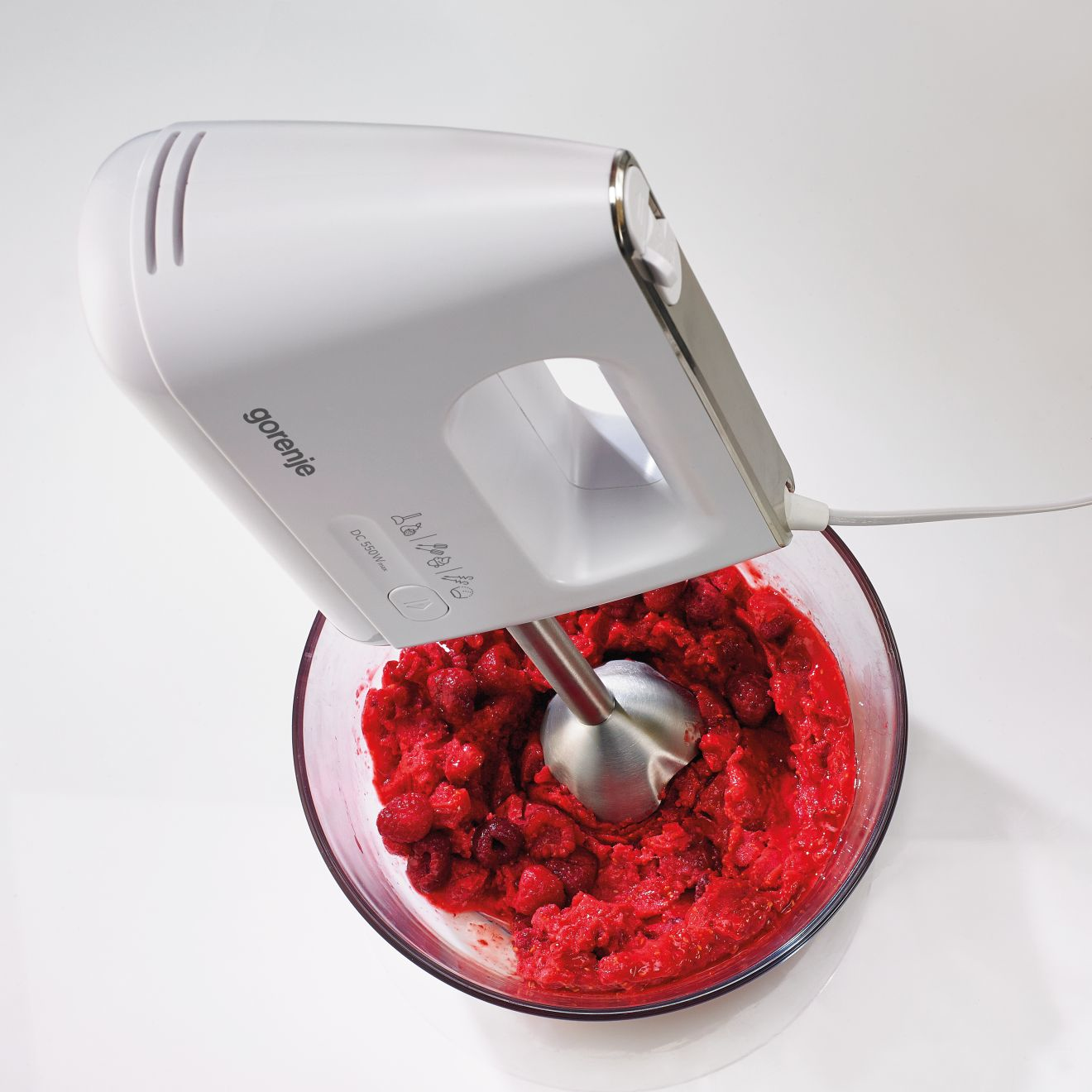 Biały blender ręczny miksuje czerwone owoce w szklanej misce na białej powierzchni.