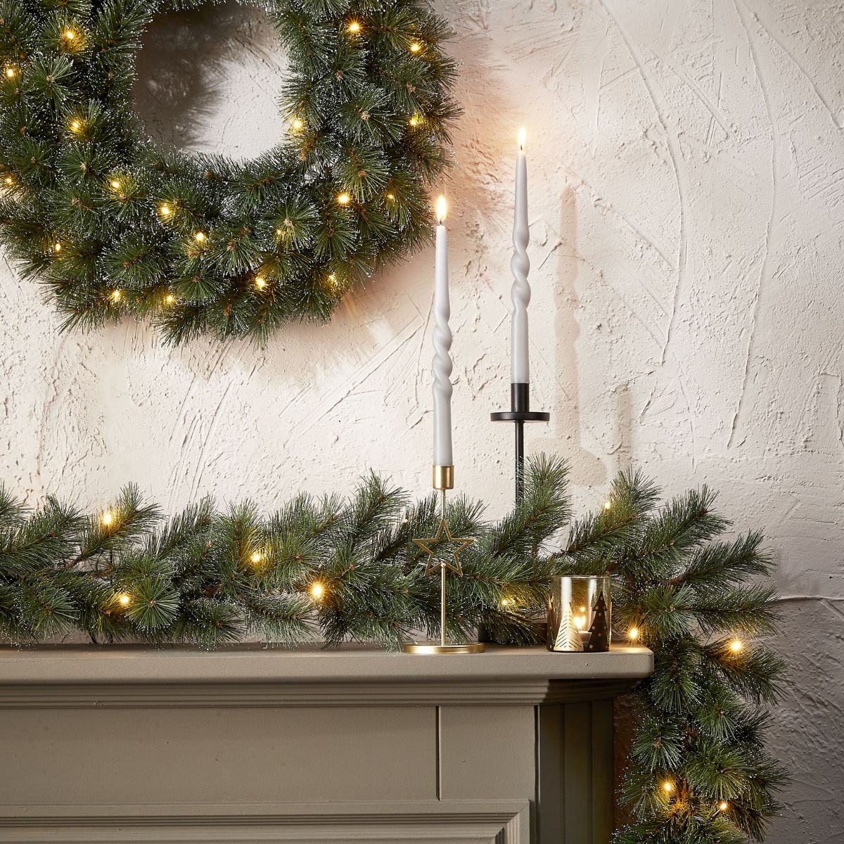 Beleuchteter Weihnachtskranz, Girlande und zwei Kerzen auf einem Kaminsims.