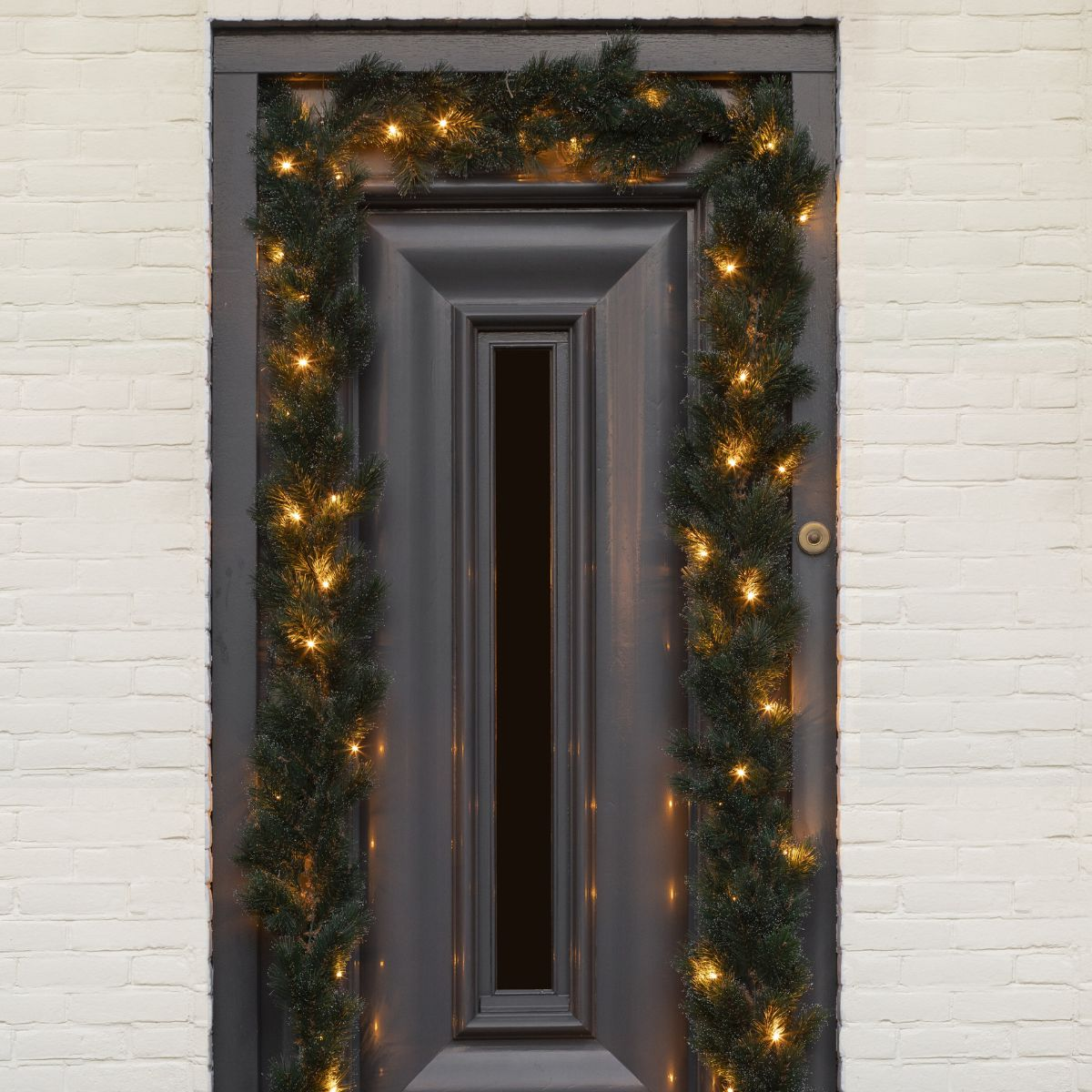 Eine graue Haustür mit einer grünen Weihnachtsgirlande und warmen Lichterketten an einer weißen Backsteinwand.