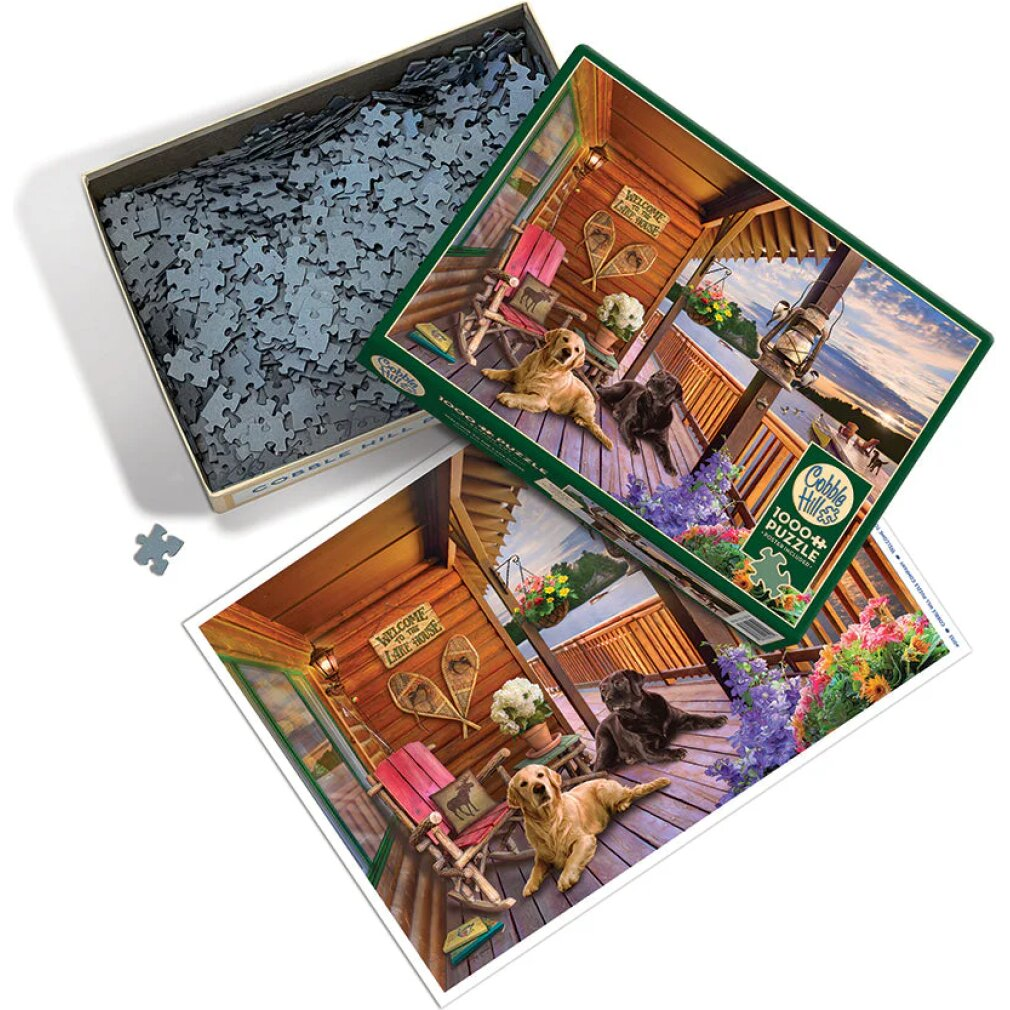 Eine offene Puzzle-Schachtel mit Teilen, Deckel und Poster zeigt eine Hüttenveranda mit Hunden bei Sonnenuntergang.