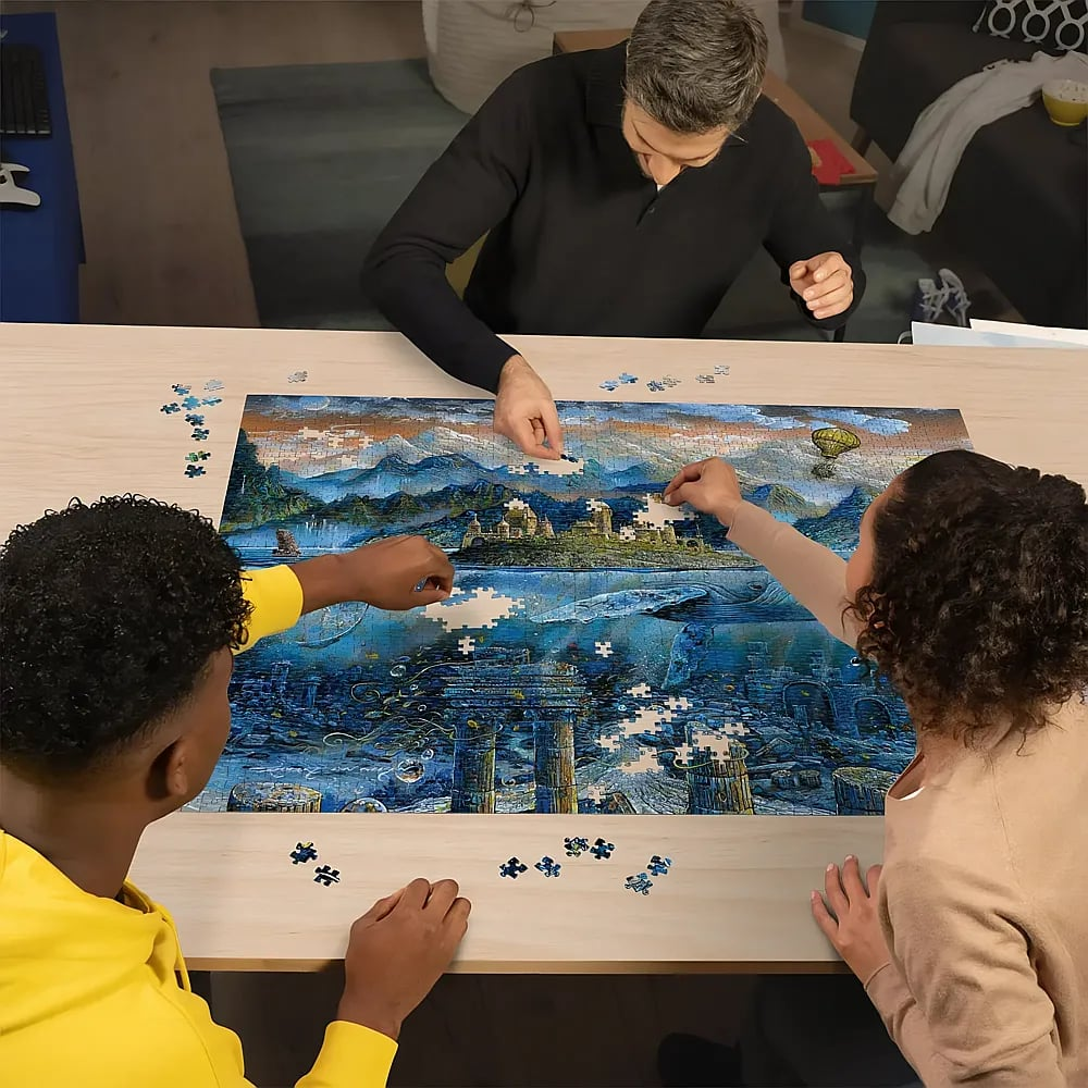 Drei Personen sitzen an einem Holztisch und setzen gemeinsam ein großes Unterwasser- und Berg-Puzzle zusammen.
