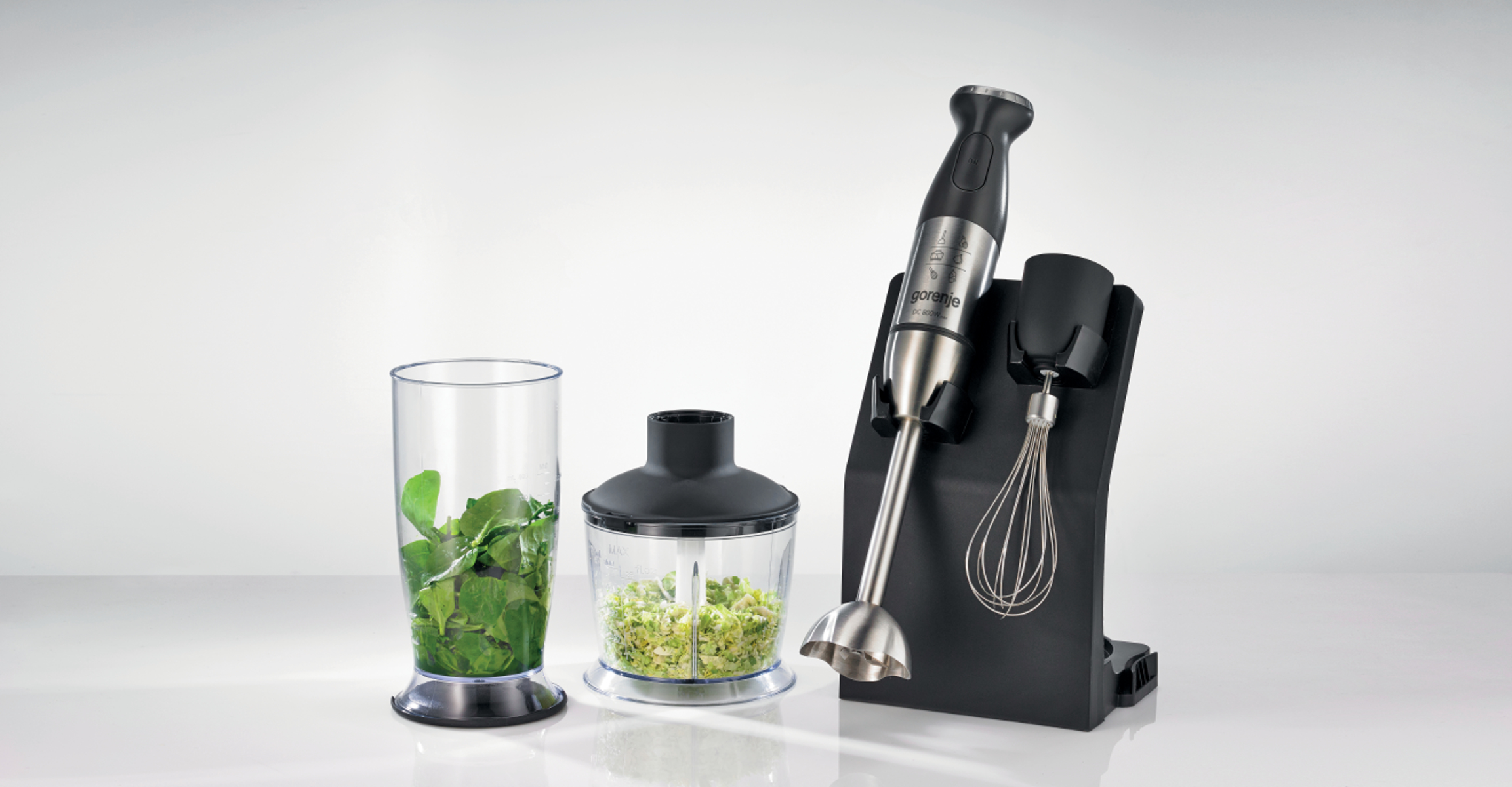 Srebrny blender ręczny z akcesoriami, w tym kubek ze szpinakiem i rozdrabniaczk na białej powierzchni.
