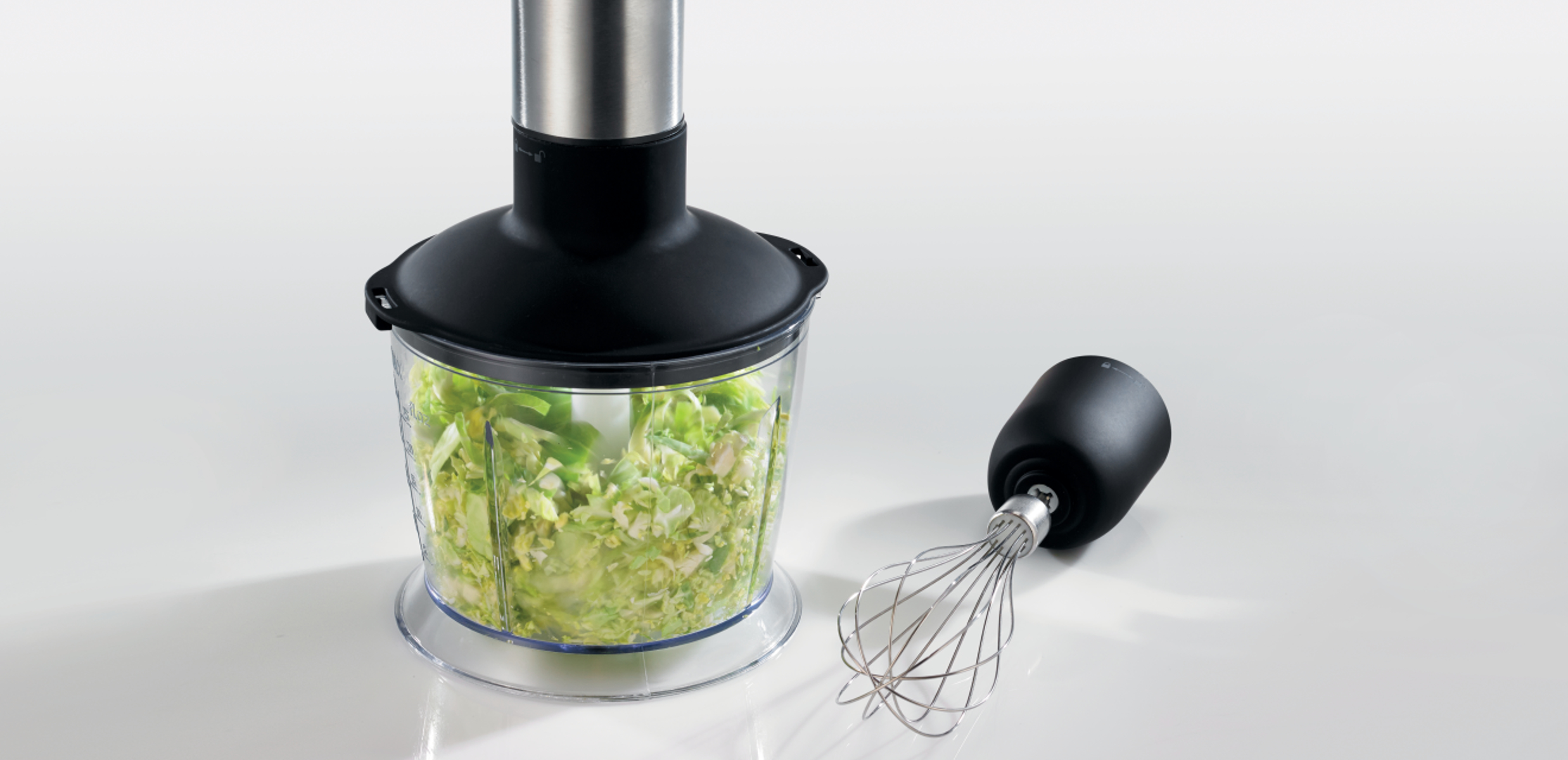 Czarny blender ręczny z pojemnikiem akrylowym wypełnionym posiekanymi warzywami i trzepaczką na białym tle.