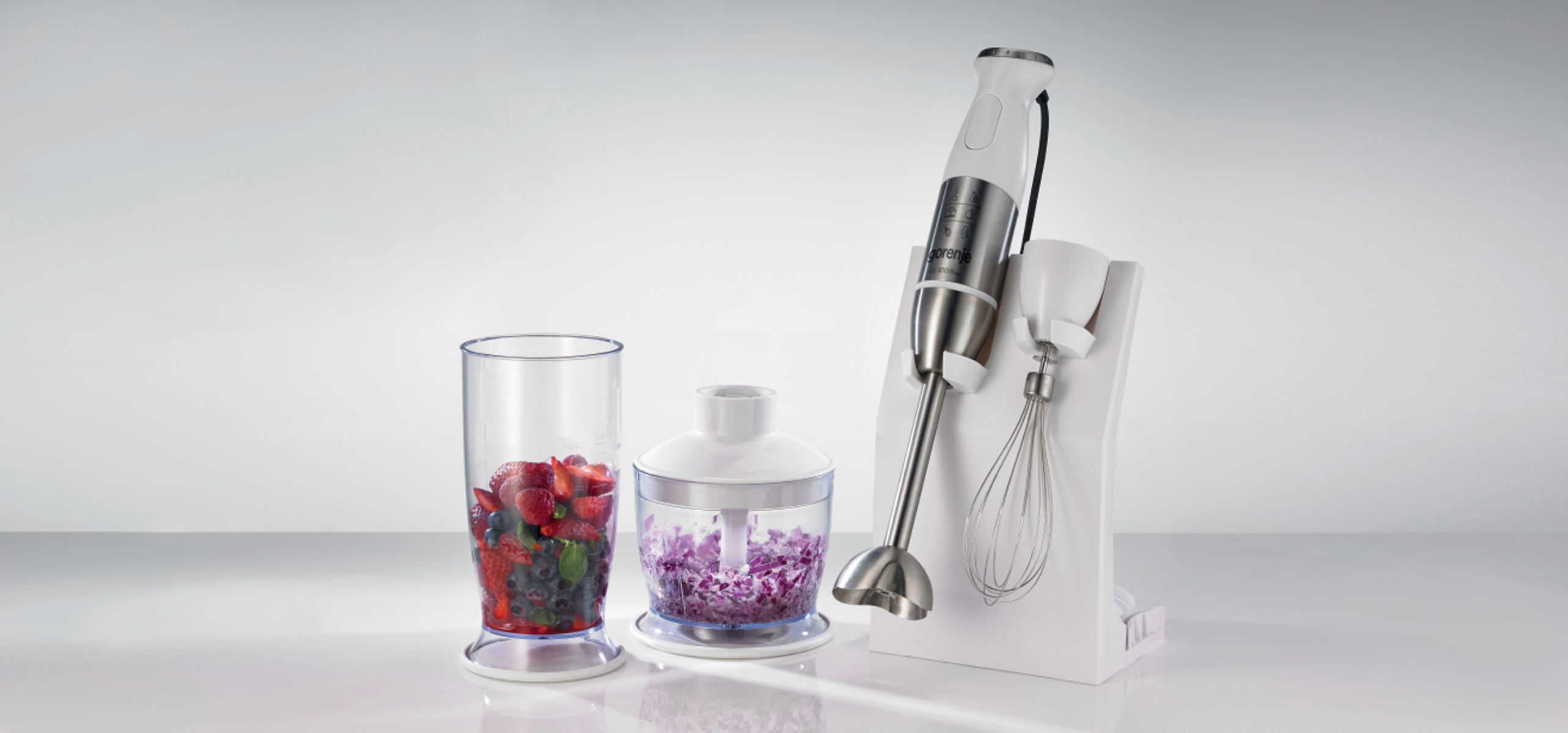 Biały blender ręczny z kubkiem, rozdrabniaczem i stojakiem na stole.