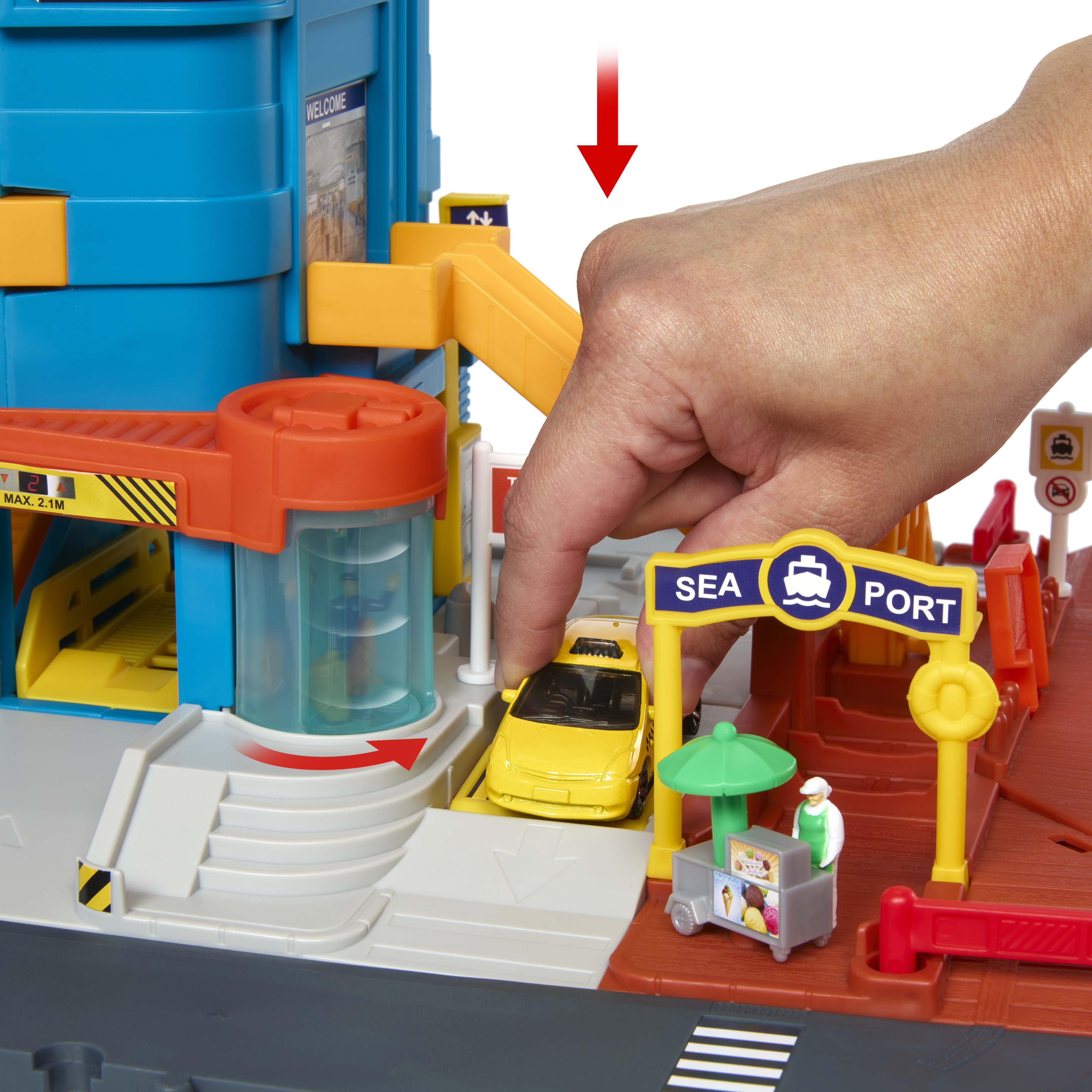 Eine Hand platziert ein gelbes Spielzeugauto in der Nähe eines Seehafen-Spielsets mit blauem Gebäude und Eimerstand.
