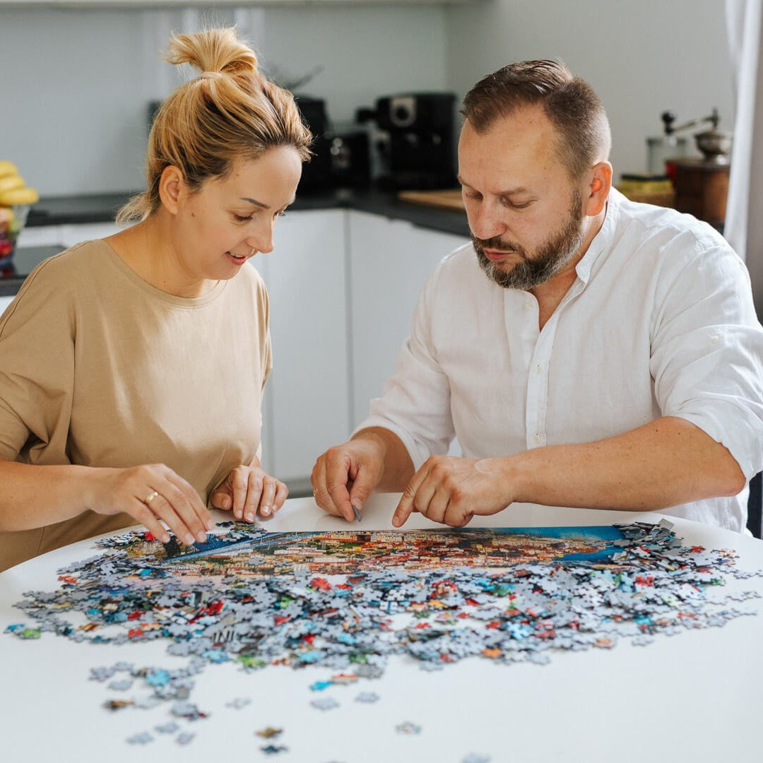 Ein Mann und eine Frau sitzen an einem weißen Tisch und setzen ein großes Puzzle zusammen.