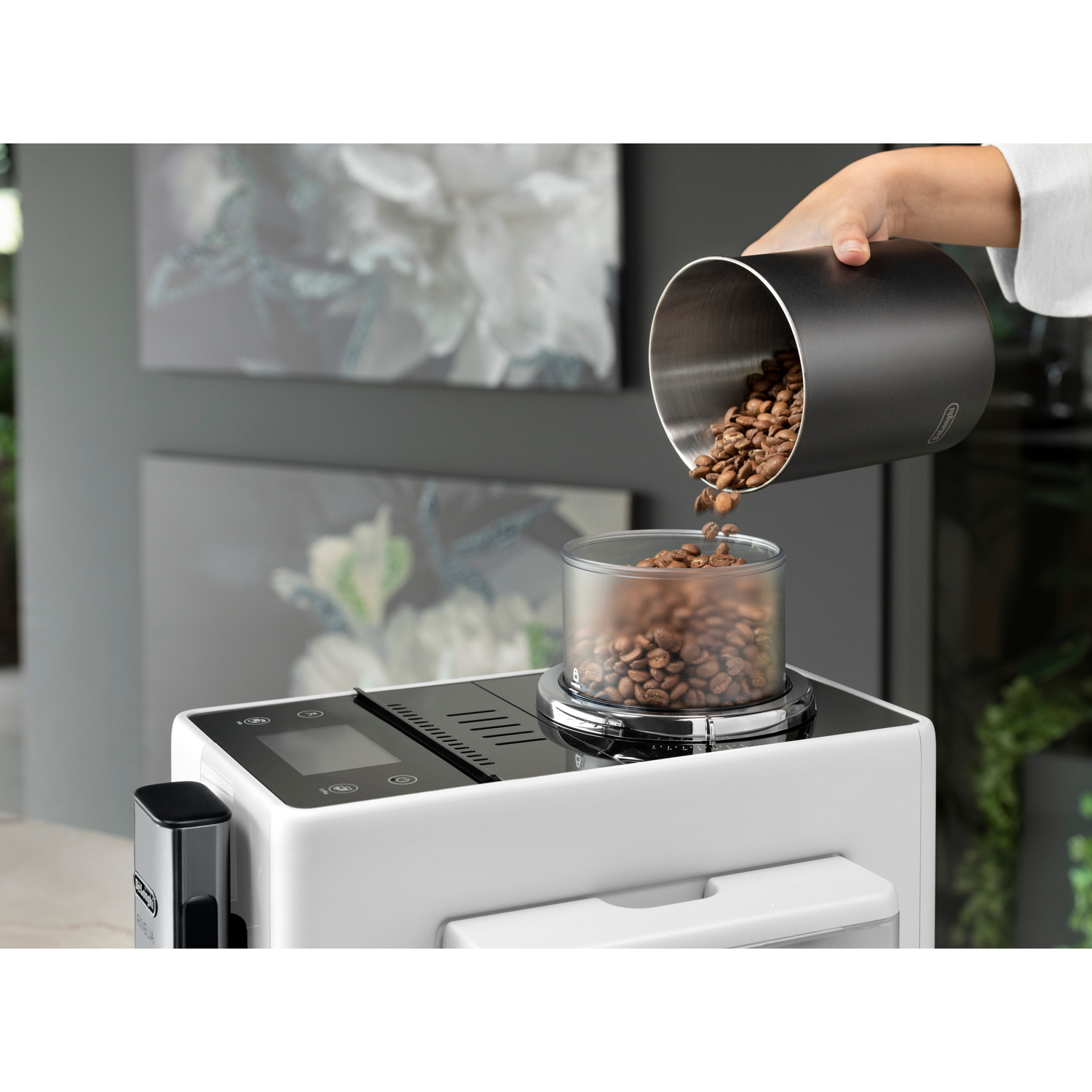 Eine Hand schüttet Kaffeebohnen aus einem Behälter in die Mühle einer weißen Kaffeemaschine.