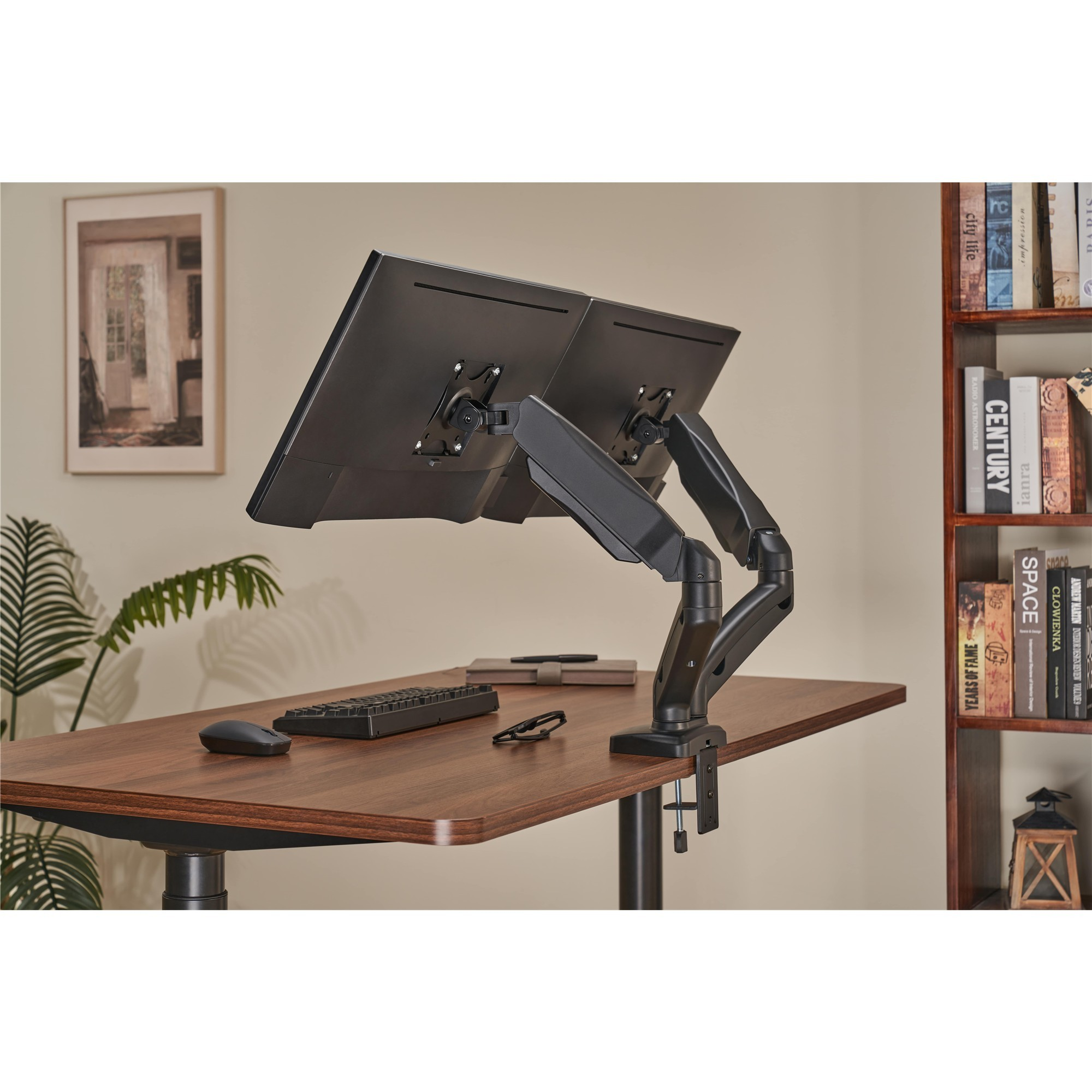 Zwei schwarze Monitorarme auf einem Holztisch mit Tastatur, Maus und Bücherregal im Hintergrund.