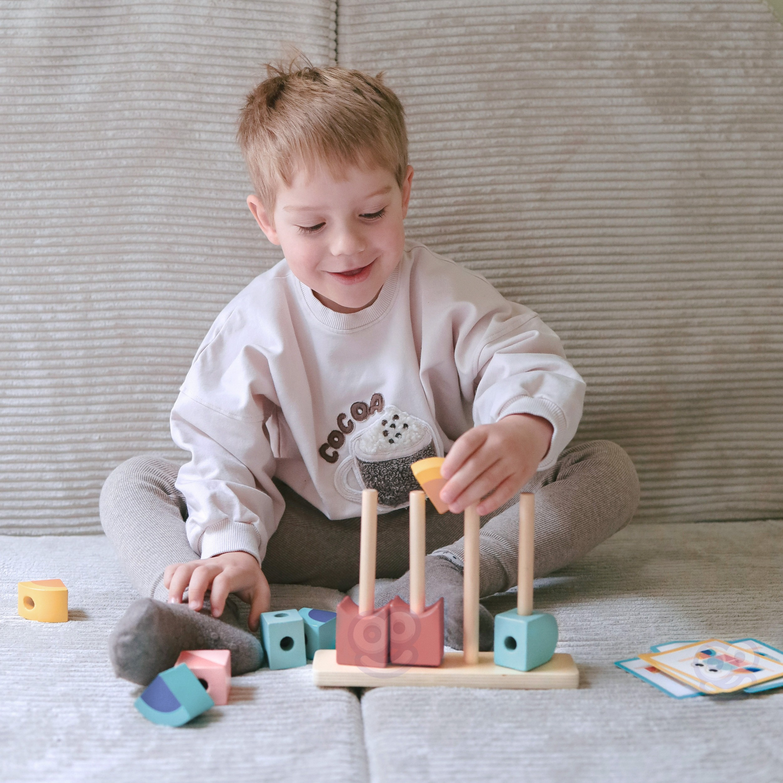 Ein kleiner Junge sitzt auf einem beigen Sofa und spielt mit einem Holzspielzeug.