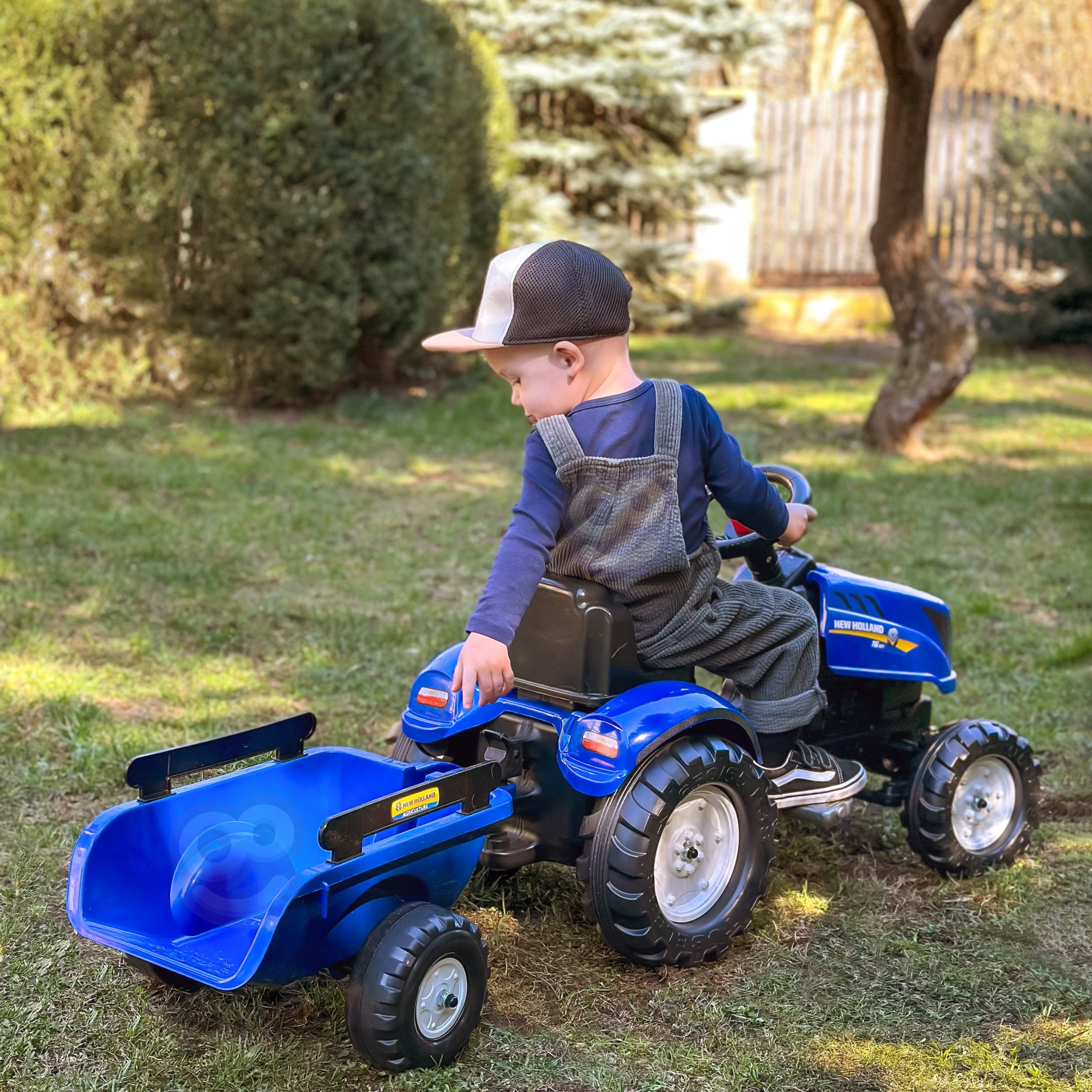 Bambino con salopette e berretto siede su un trattore giocattolo blu con rimorchio in un giardino.