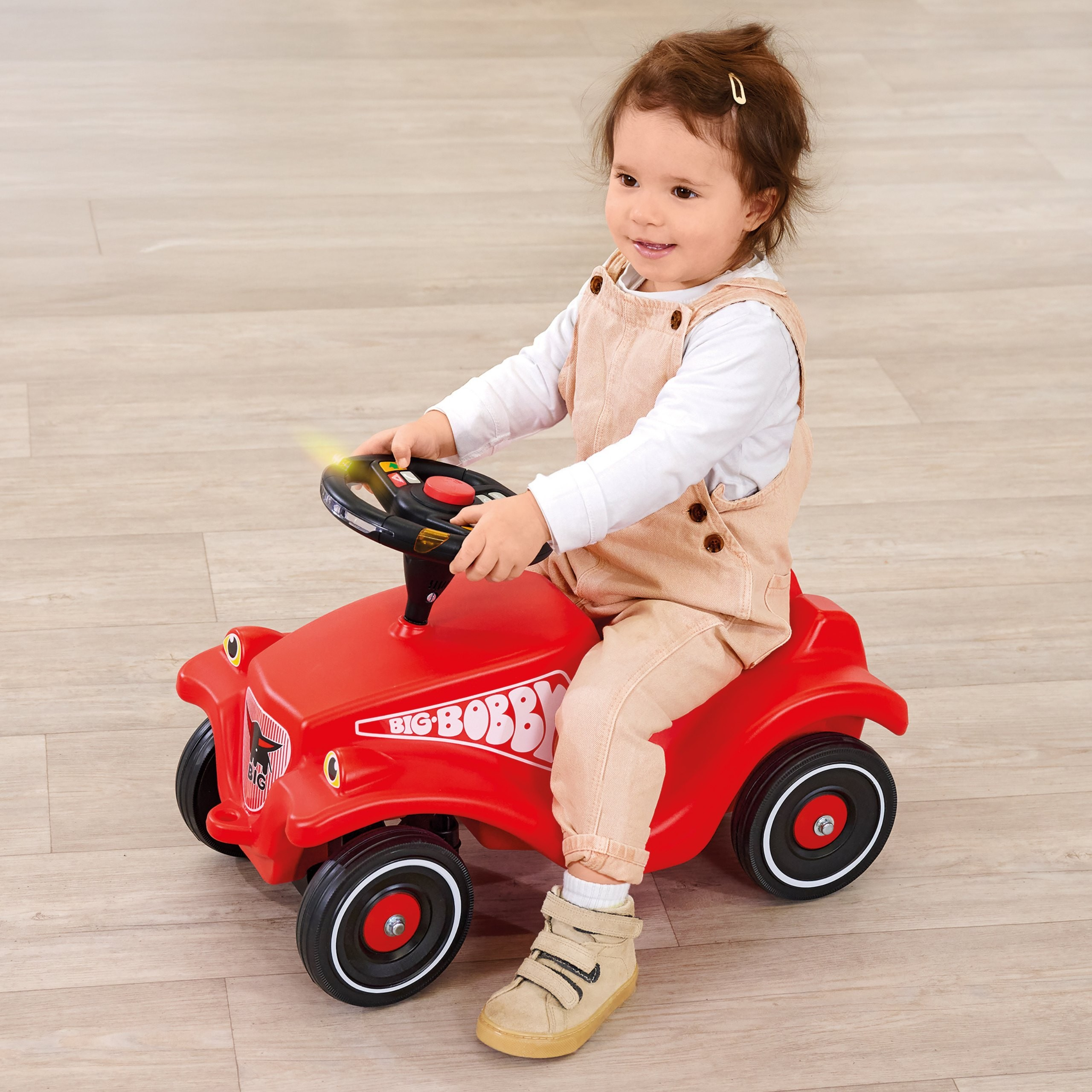 Ein lächelndes Kleinkind fährt auf einem Holzboden ein hellrotes Spielzeugauto.