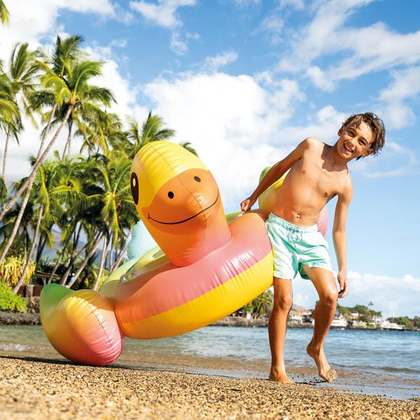 Ein Junge mit Badehose zieht eine große, bunte Enten-Schwimminsel über den Sandstrand am tropischen Meer.