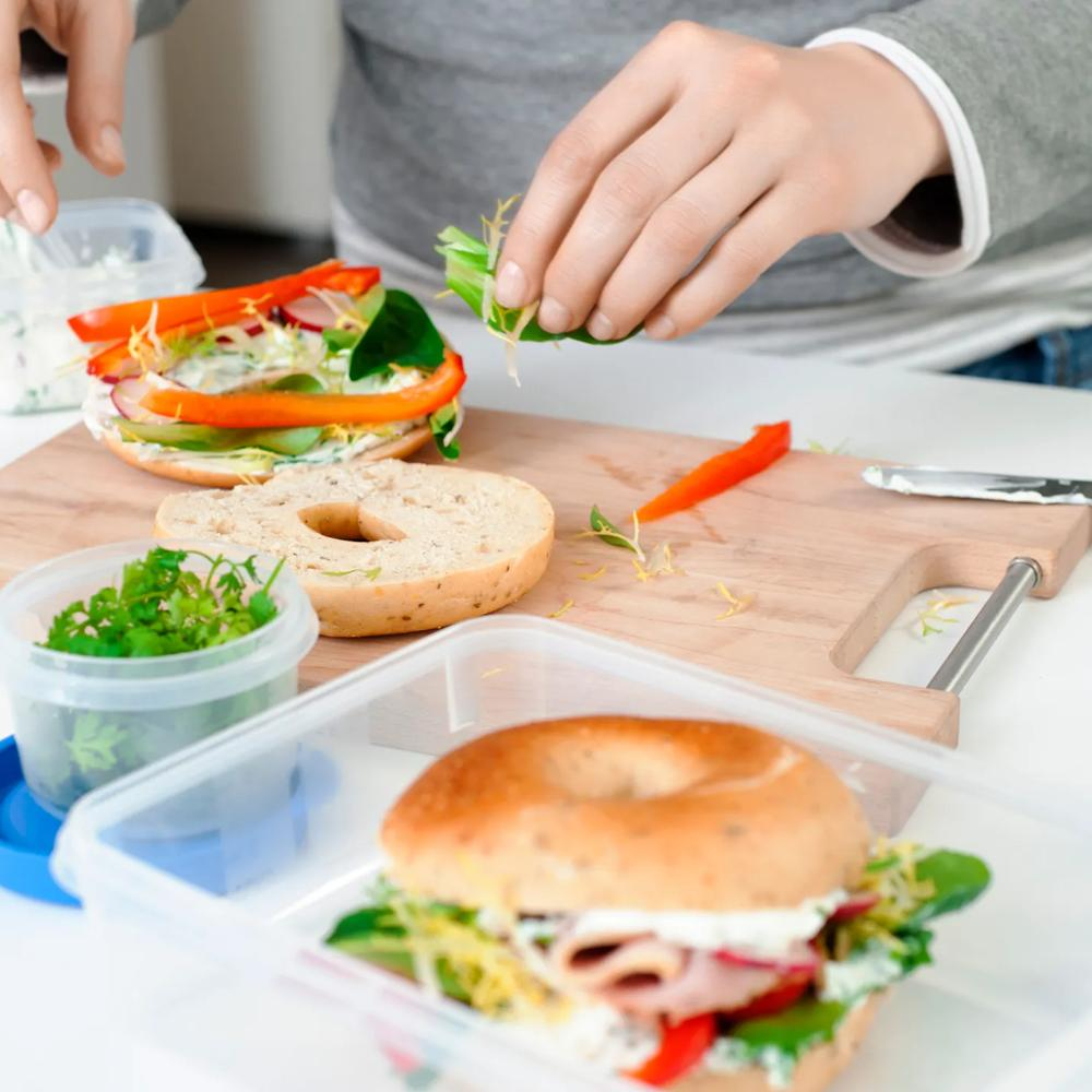 Eine Person bereitet ein Bagelsandwich mit Gemüse und Aufstrich auf einem Holzschneidebrett zu.