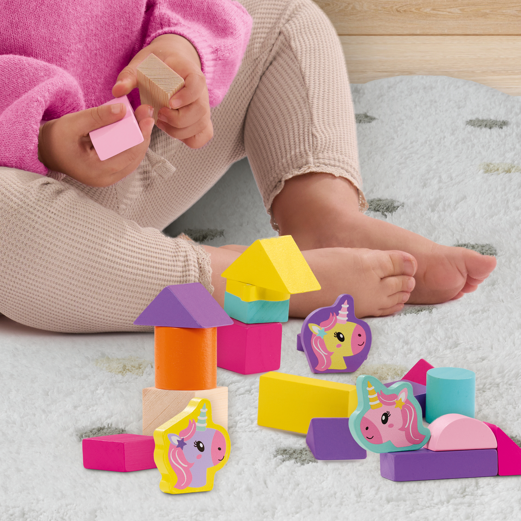 Ein Kind in rosa Kleidung sitzt auf einem Teppich und spielt mit bunten Holzbauklötzen und Einhornfiguren.
