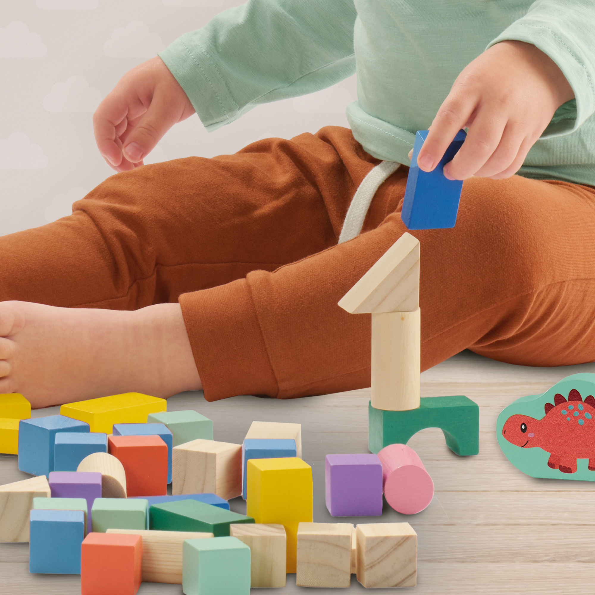 Kleinkind in grünem Oberteil und braunen Hosen spielt auf dem Boden mit bunten Holzbauklötzen.