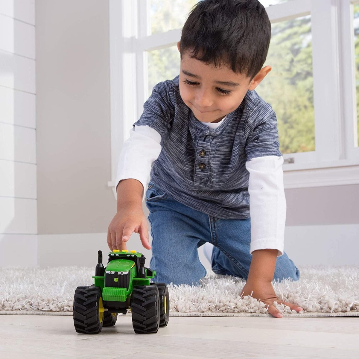Un jeune garçon s'agenouille sur un tapis blanc en jouant avec un tracteur jouet vert.