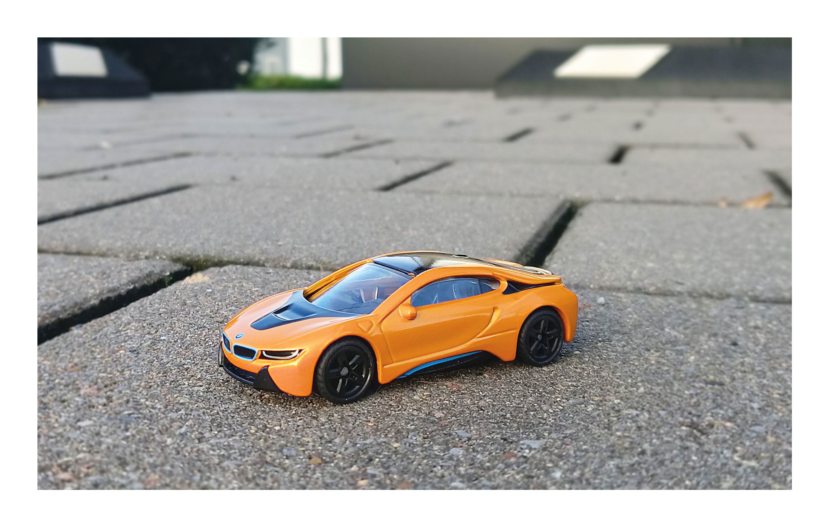 Ein orangefarbenes Spielzeugauto mit schwarzen Details steht auf einem grauen gepflasterten Boden im Freien.