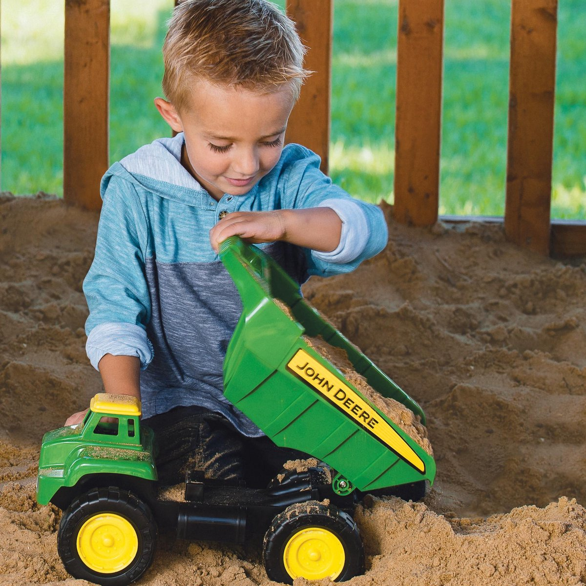 Kleiner Junge in blauem Pullover spielt im Sandkasten mit einem grünen John Deere Spielzeugkipper.
