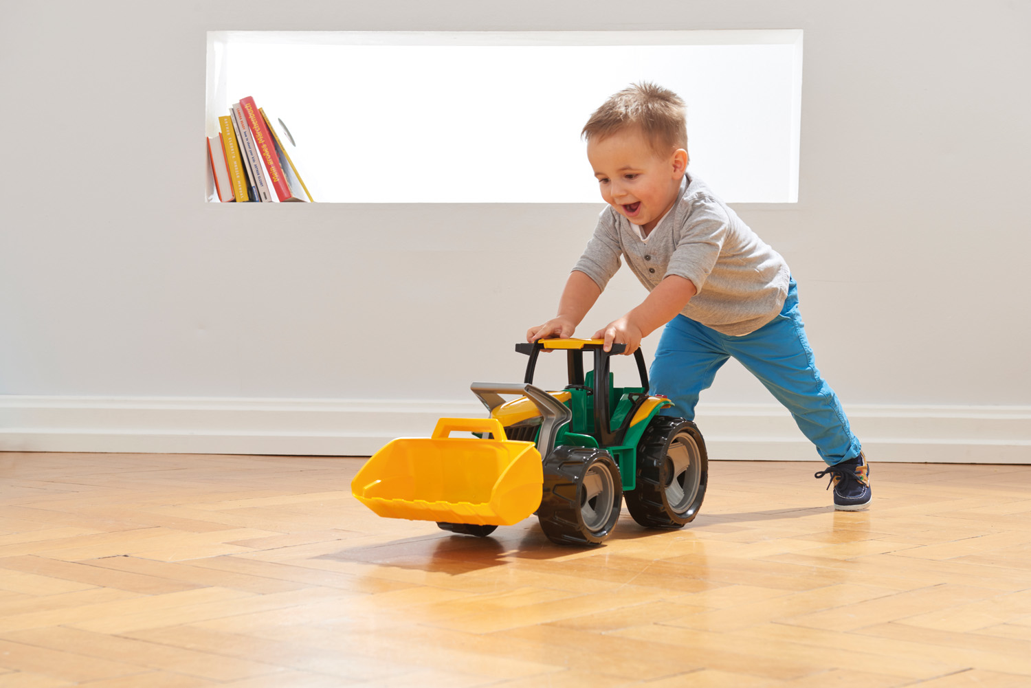 Kleinkind schiebt einen großen grünen und gelben Spielzeugtraktor mit Frontlader auf einem Holzboden.