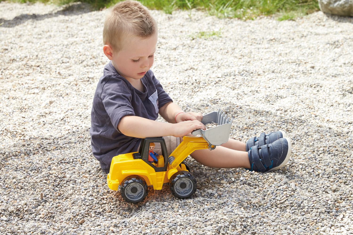 Bambino seduto all'aperto sulla ghiaia che gioca con un escavatore giocattolo giallo.