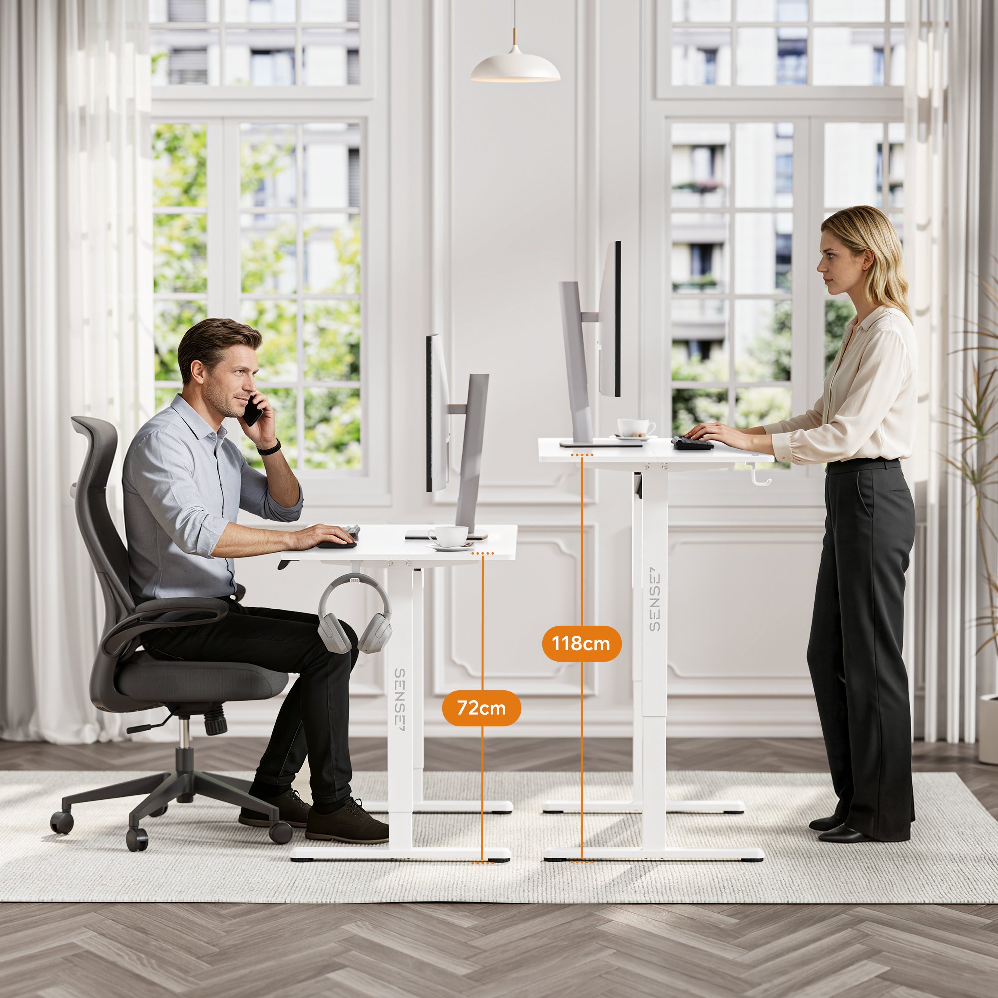 A man sits at a low desk on a phone call while a woman stands working at a high desk in a bright room.