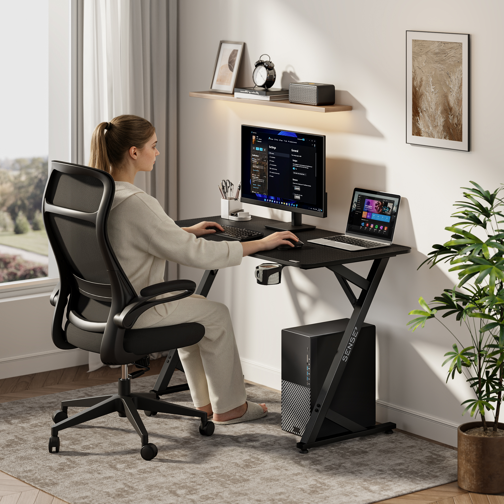 Eine Frau sitzt an einem Schreibtisch mit PC und Laptop in einem hellen Zimmer.