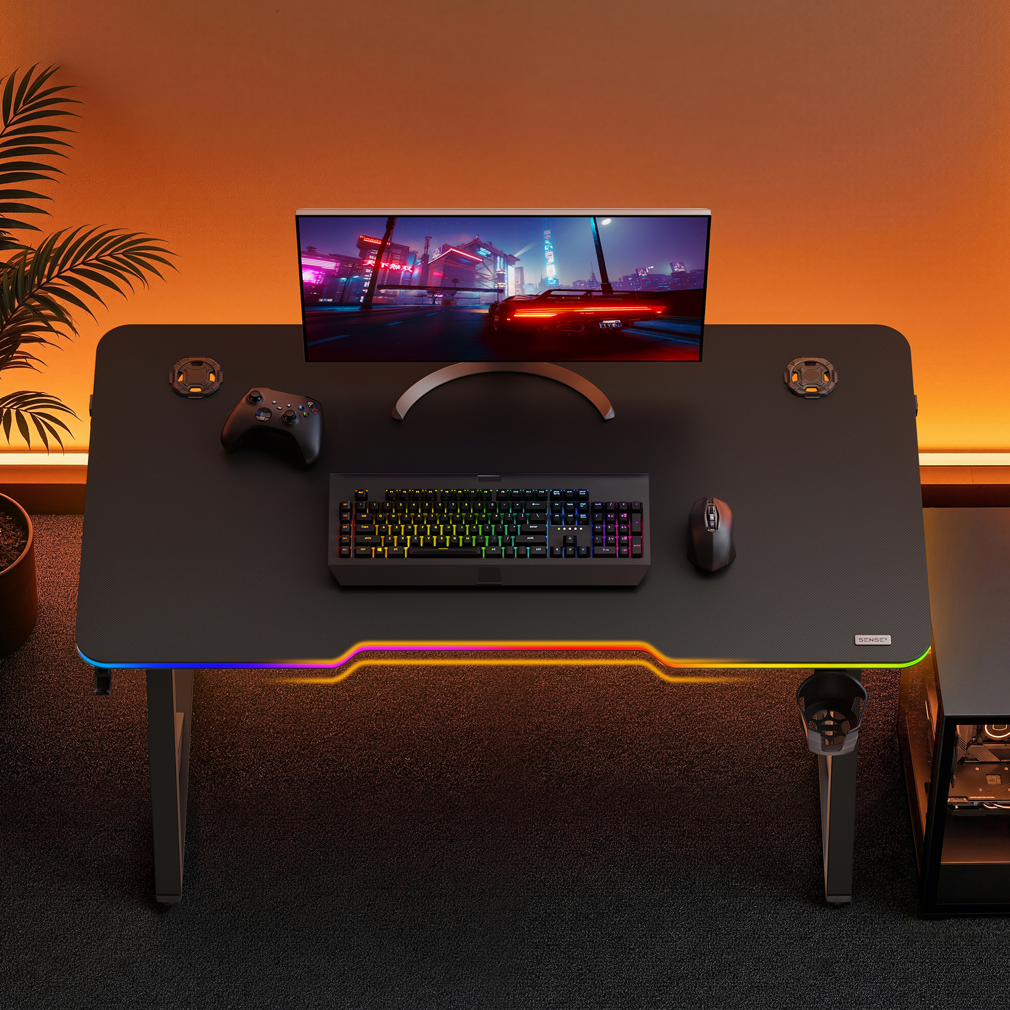 Ein Gaming-Schreibtisch mit RGB-Beleuchtung, Monitor, Tastatur, Maus und Controller vor einer orangefarbenen Wand.