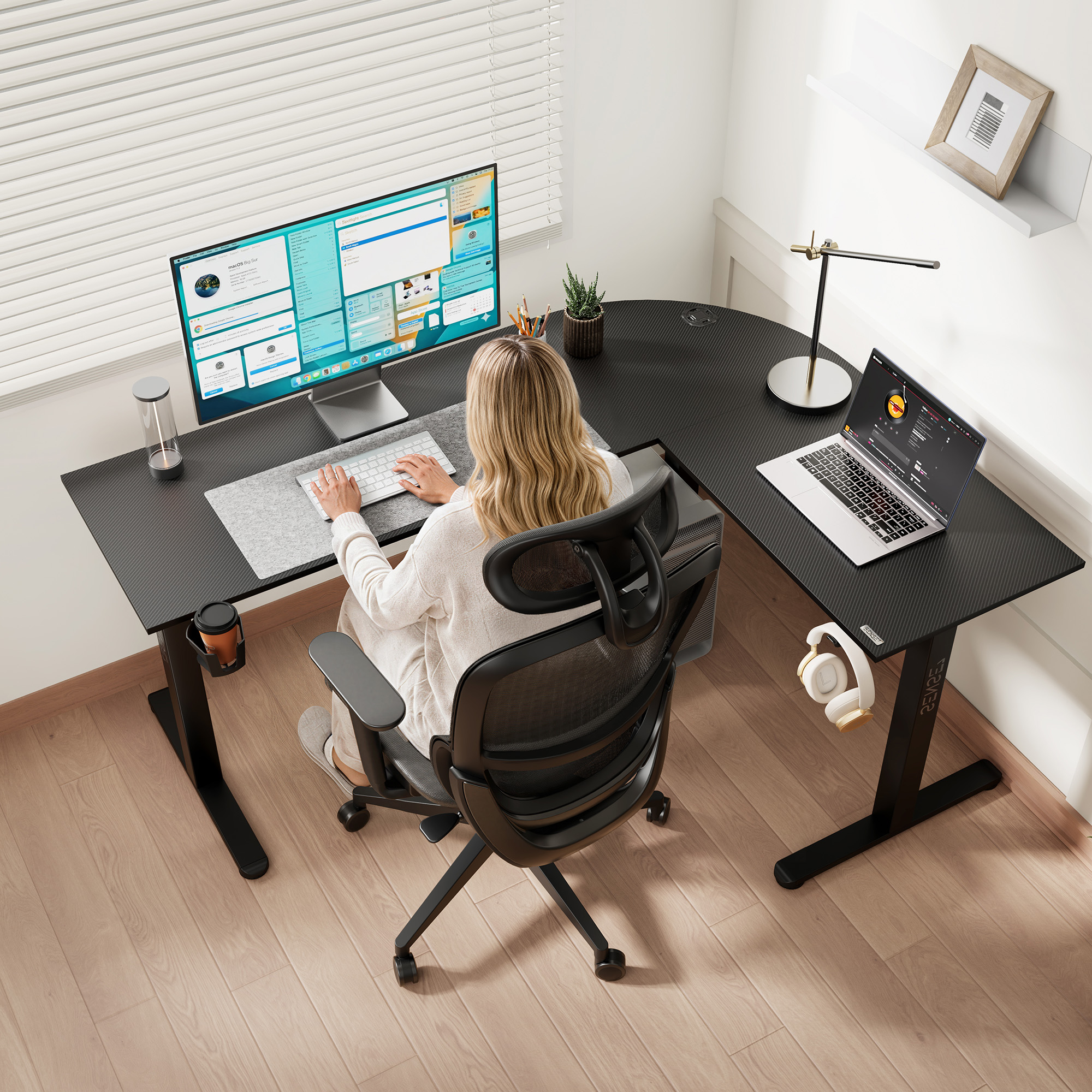 A woman works at a large L-shaped desk with a monitor, laptop, and modern accessories in a bright room.