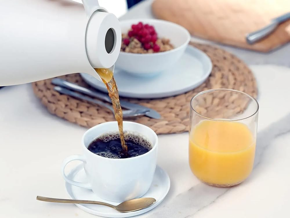 Kaffee wird aus einer Kanne in eine weiße Tasse gegossen, daneben stehen Saft und Müsli.