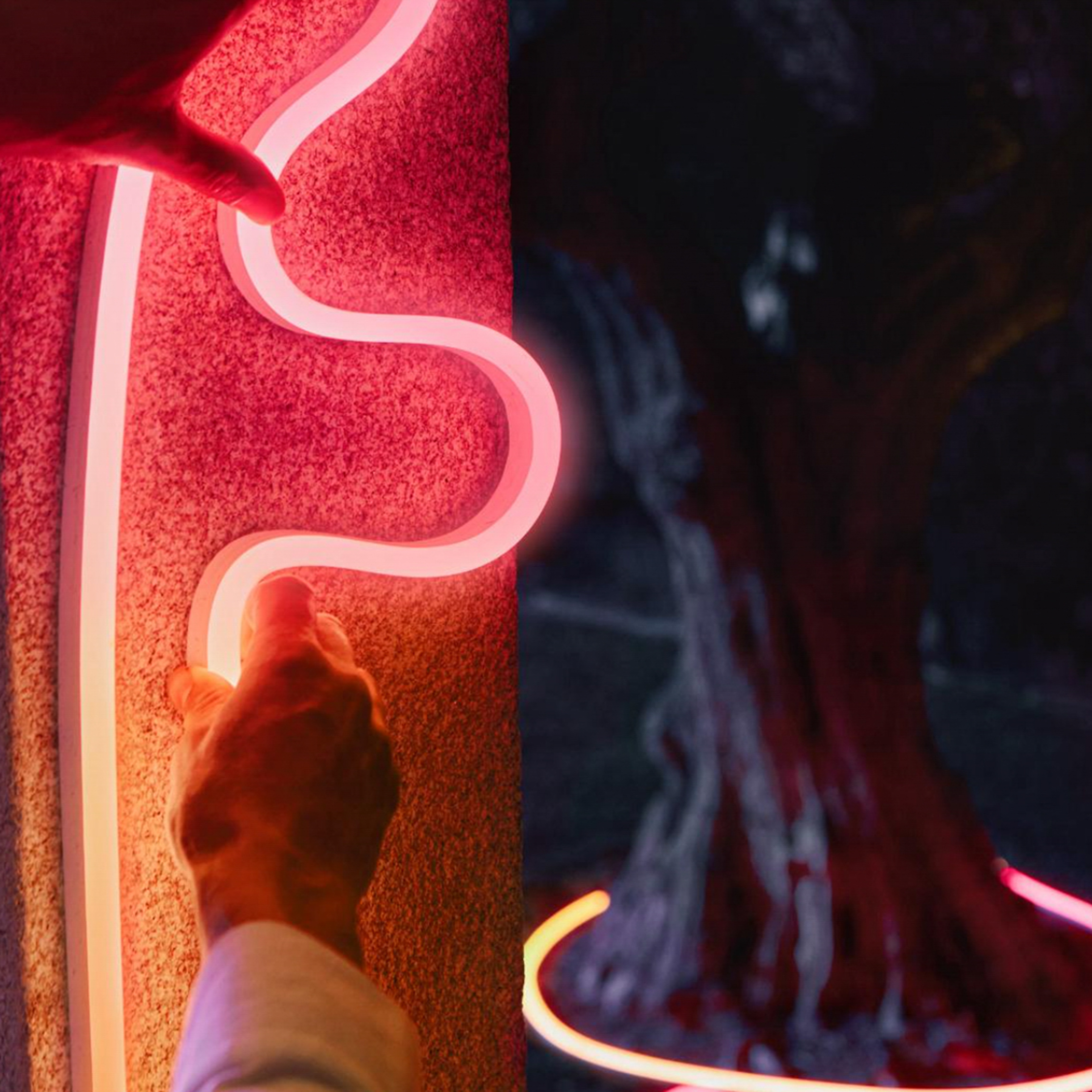 Mani posizionano una striscia LED rossa ondulata su un pilastro di pietra; sullo sfondo un albero illuminato.