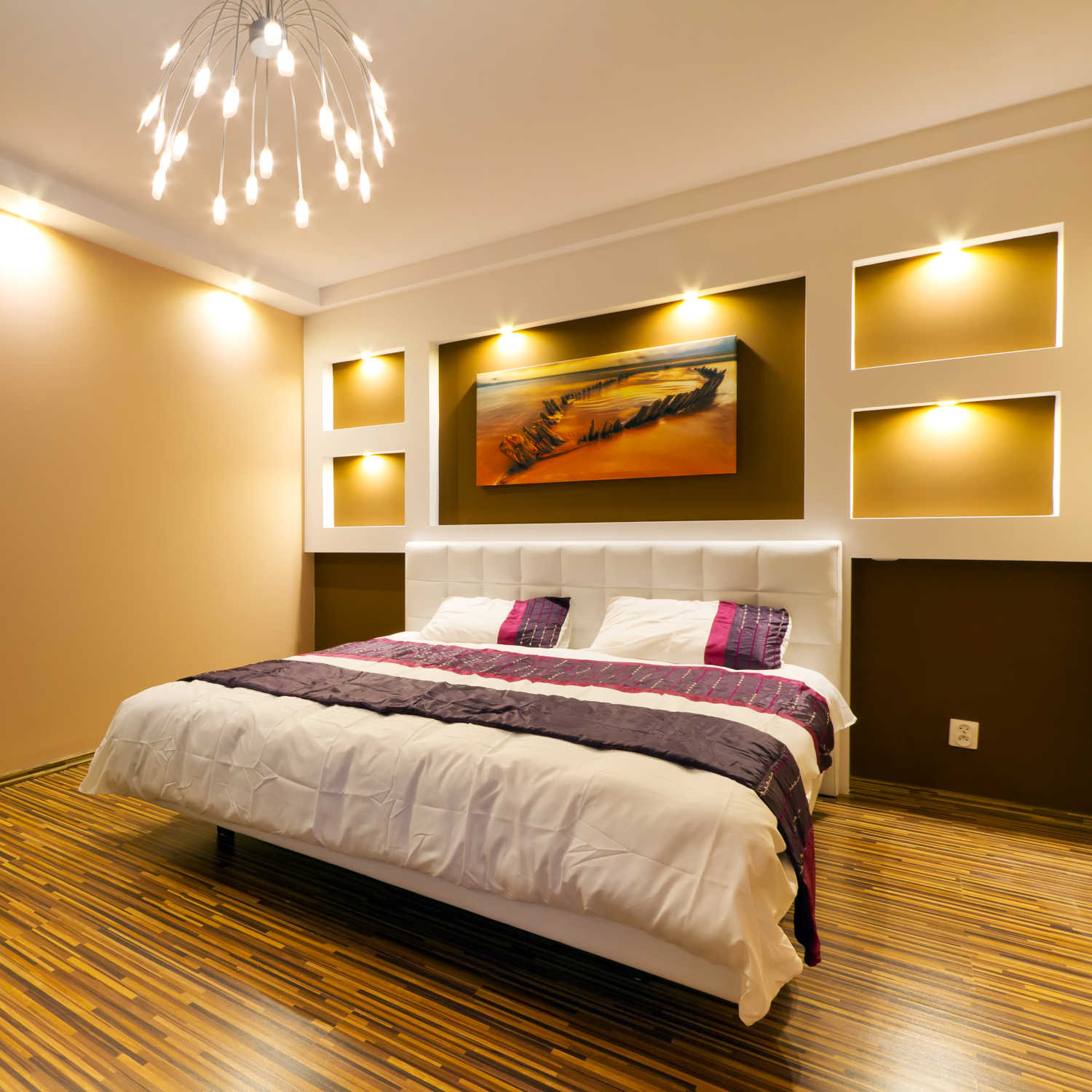 Moderna camera da letto con letto bianco, dettagli viola, nicchie illuminate, un dipinto e un lampadario unico.