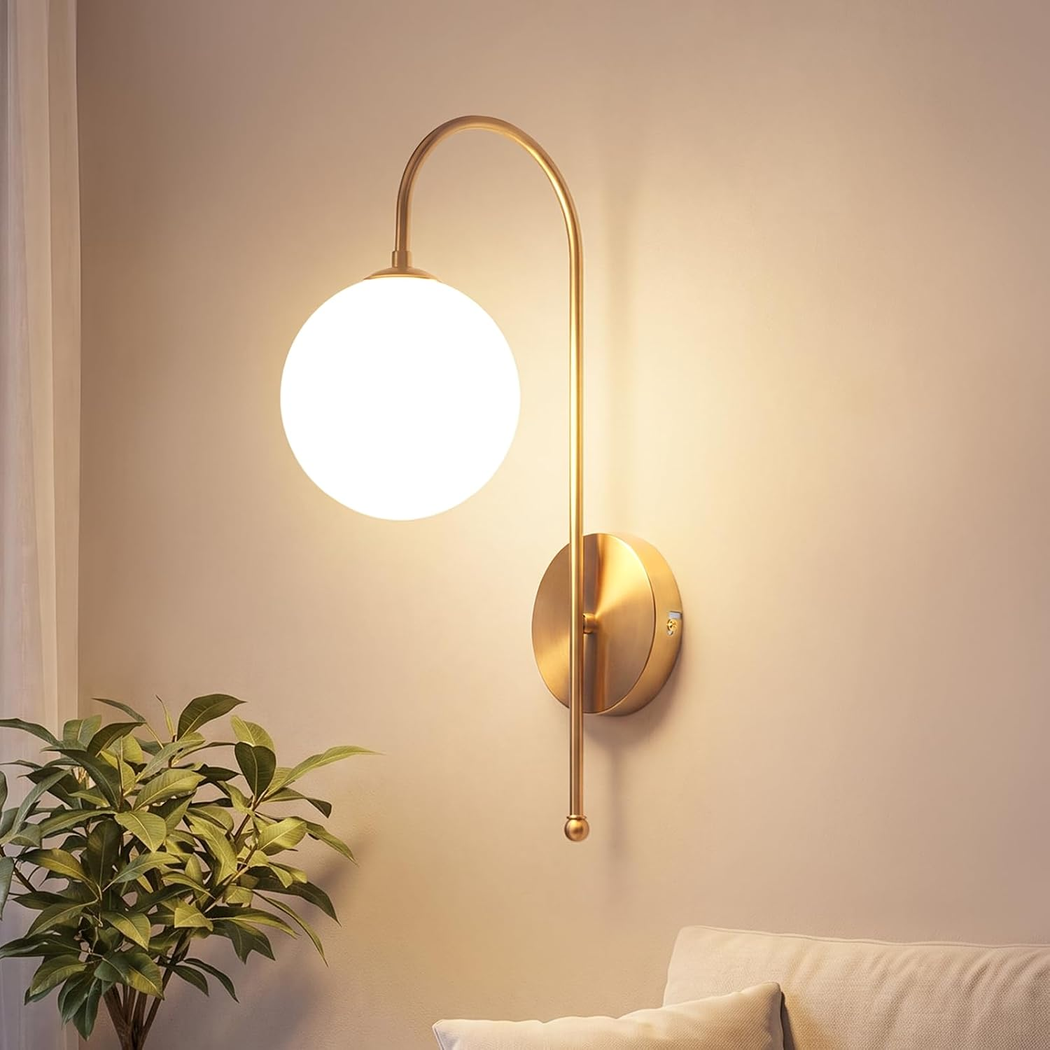 Eine goldene Wandlampe mit weißem Kugelschirm leuchtet an einer Wand neben einer Pflanze und einem Sofa.