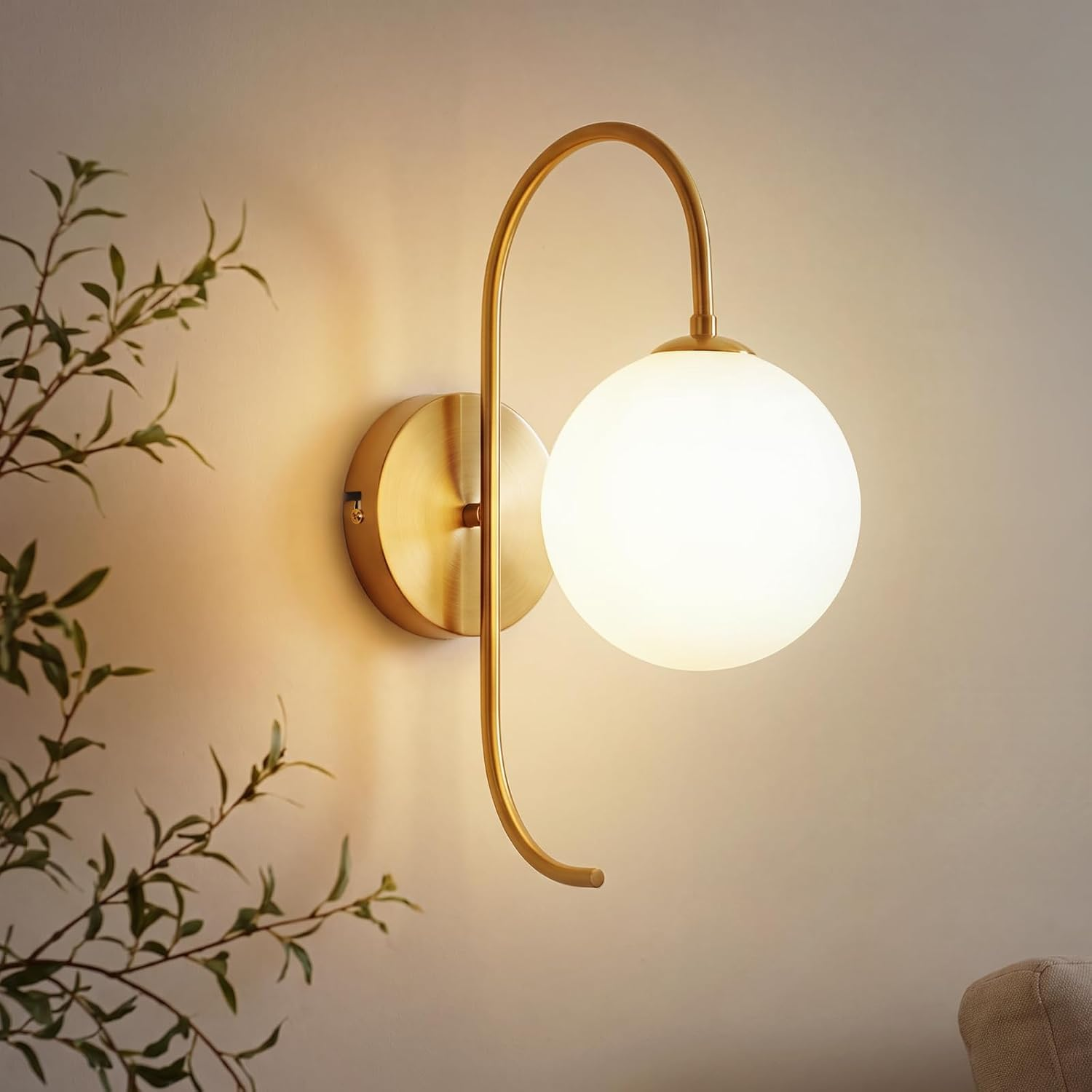 Eine goldene Wandleuchte mit einer runden, weiß leuchtenden Glaskugel hängt an einer hellen Wand neben Pflanzen.