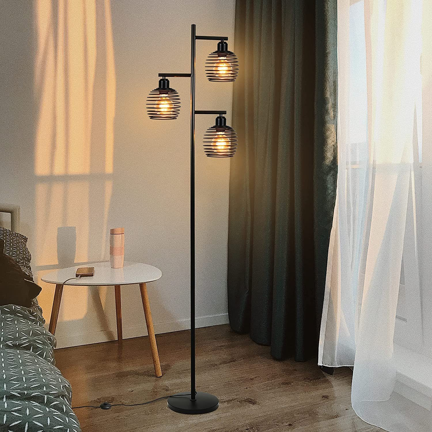 Eine schwarze Stehlampe mit drei Glühbirnen im Käfigdesign steht in einem Schlafzimmer neben einem Bett und Vorhängen.