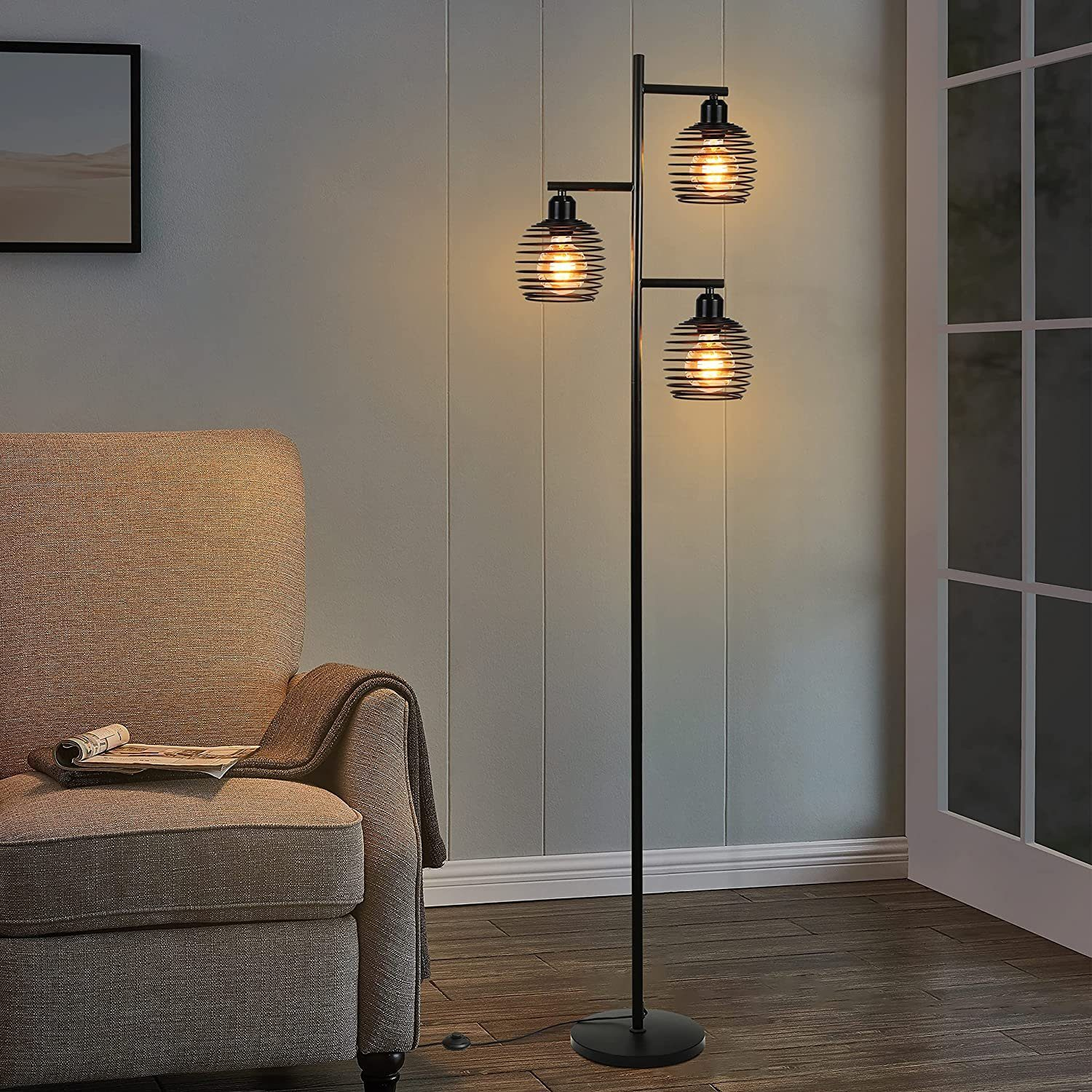 Eine schwarze Stehlampe aus Metall mit drei Drahtschirmen steht in einem Wohnzimmer neben einem Sessel.
