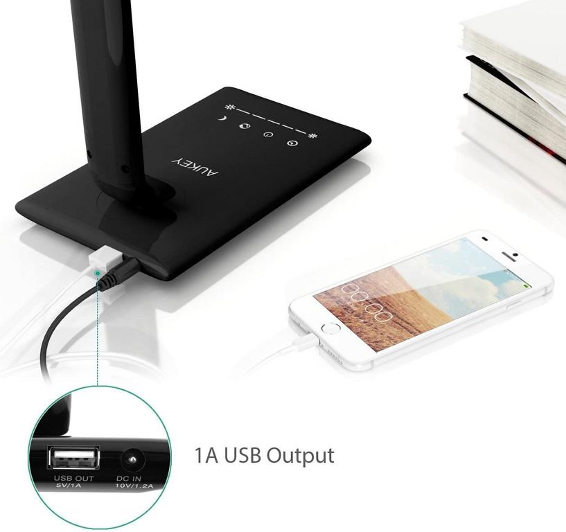 Ein schwarzer Schreibtischlampensockel mit Touch-Steuerung, eingestecktem USB-Kabel, ladendem weißen Smartphone und Büchern.