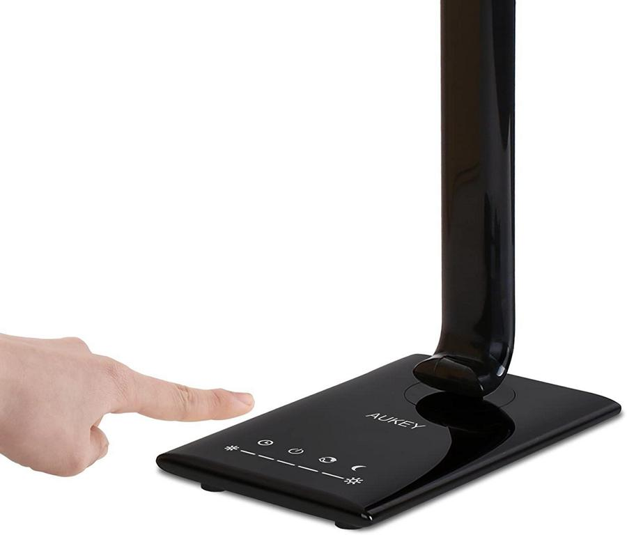 Eine Hand berührt das Touch-Bedienfeld einer glänzenden schwarzen Aukey-Schreibtischlampe.
