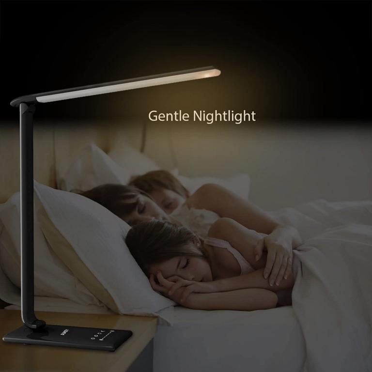 Eine schwarze Schreibtischlampe leuchtet sanft auf einem Nachttisch, während zwei Personen im Bett schlafen.