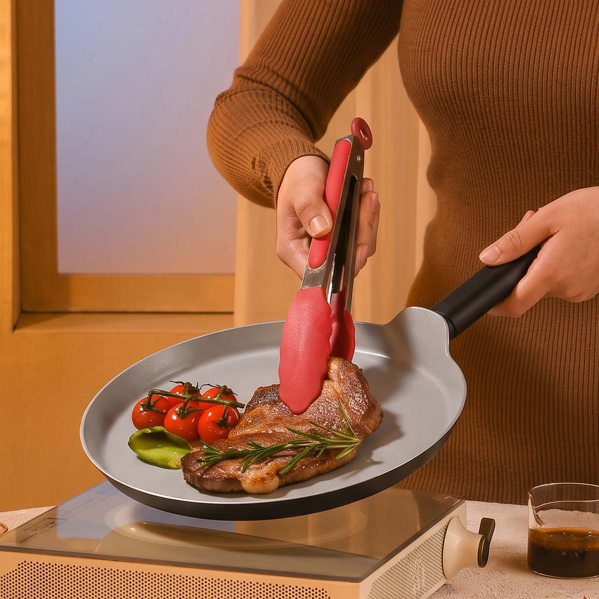Una persona sostiene pinzas rojas y voltea un bistec con tomates cherry en una sartén.