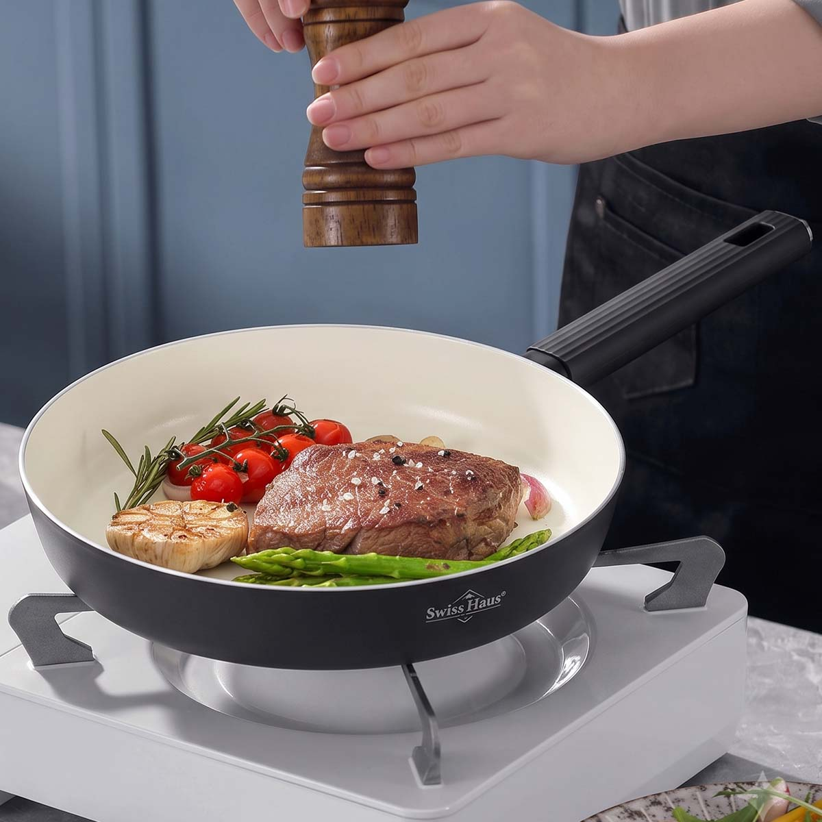 Una persona sazona bistec, espárragos, tomates y ajo con pimienta en una sartén Swiss Haus.