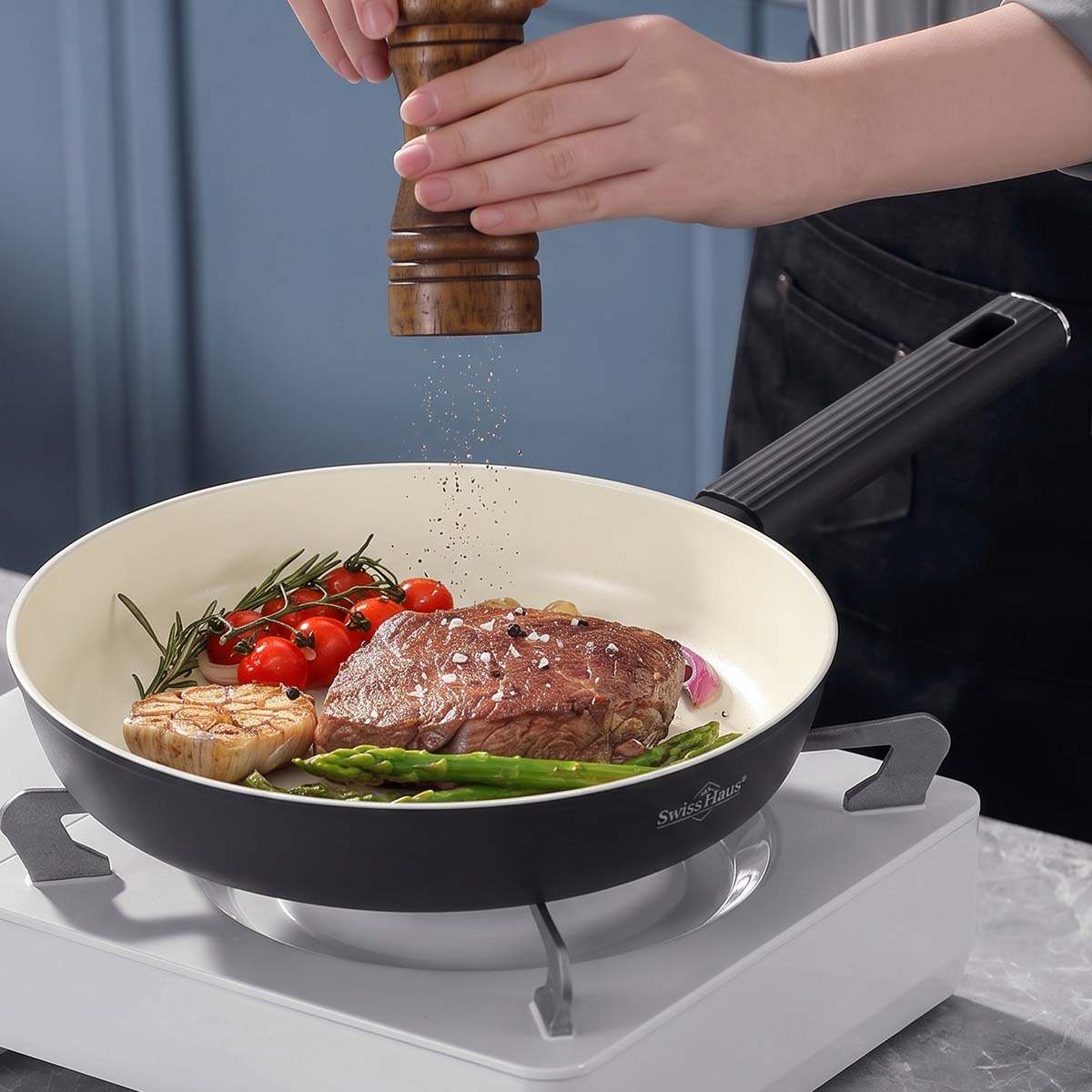Alguien muele pimienta sobre un bistec con espárragos y tomates en una sartén.