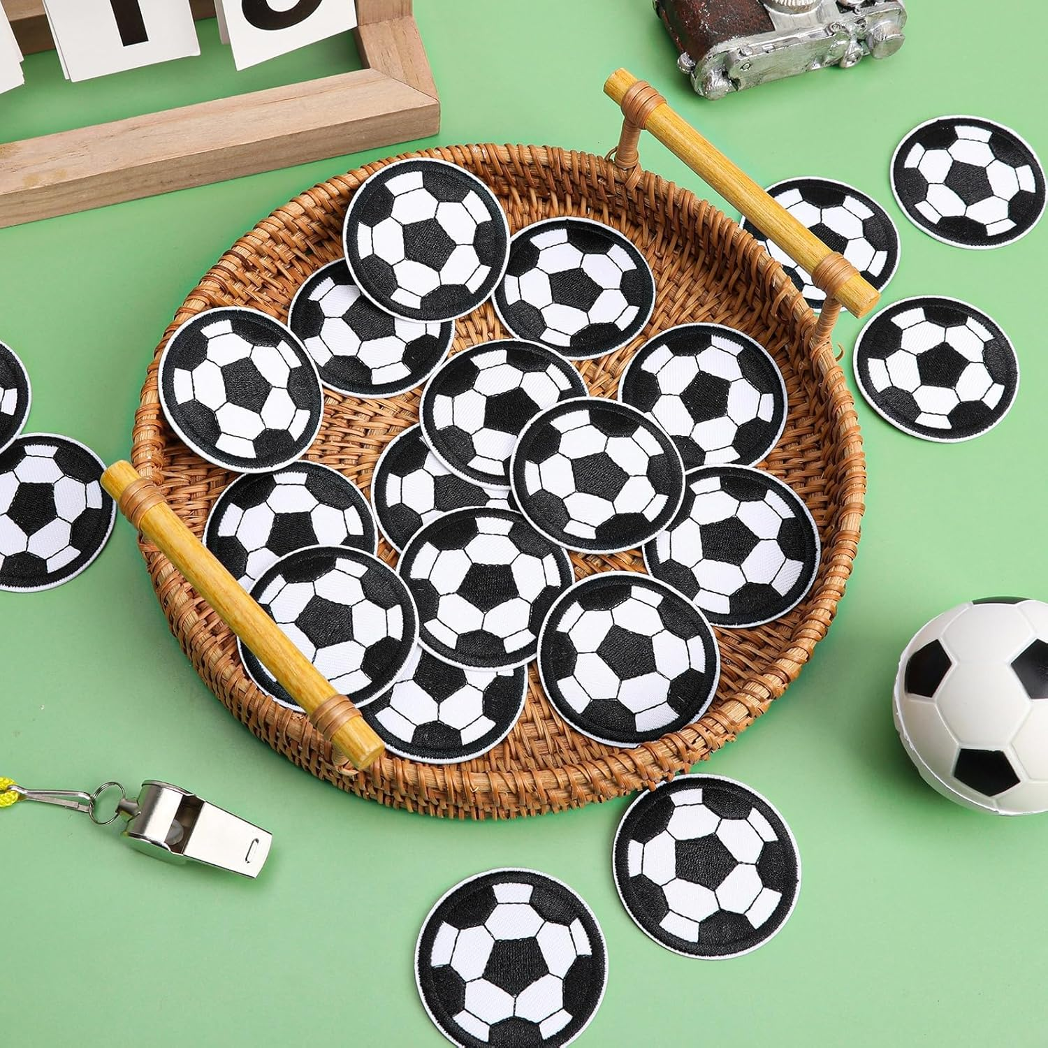 Viele schwarz-weiße Fußball-Aufnäher in einem geflochtenen Tablett mit einem Fußball und einer Pfeife auf grünem Grund.