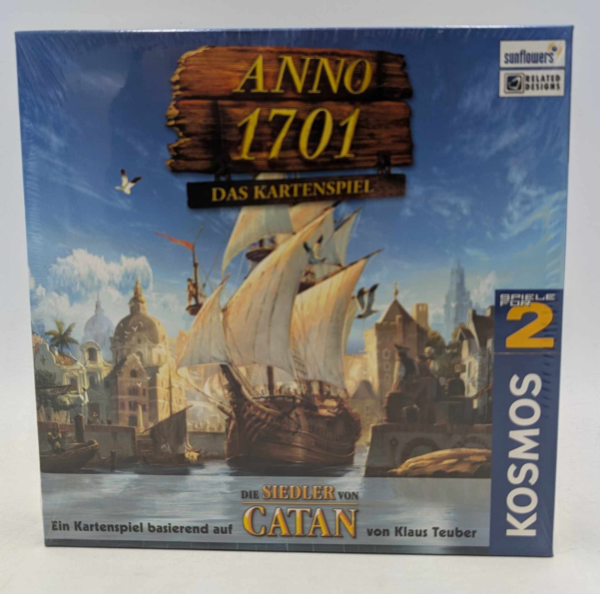 Anno 1701 Kartenspiel-Schachtel zeigt ein Segelschiff im Hafen mit Stadt im Hintergrund und Holzschild.