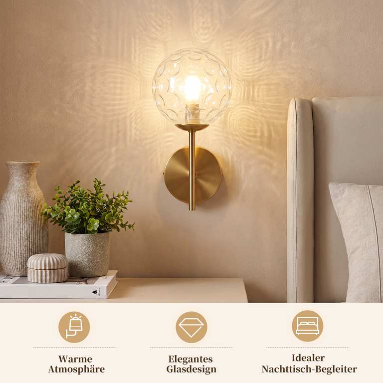 Eine goldene Wandlampe mit Glaskugel strahlt warmes Licht auf die Wand, neben einem Bett.
