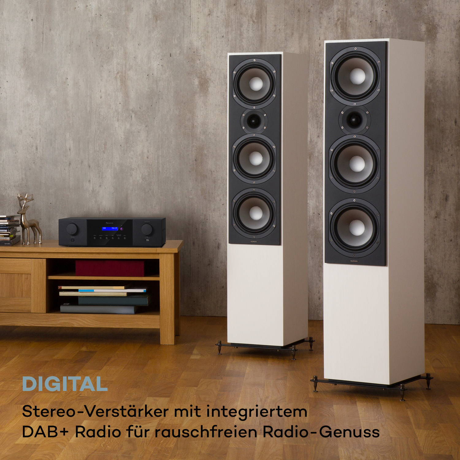 Zwei weiße Standlautsprecher und ein schwarzer Stereo-Verstärker stehen in einem Raum mit Holzboden und Betonwand.